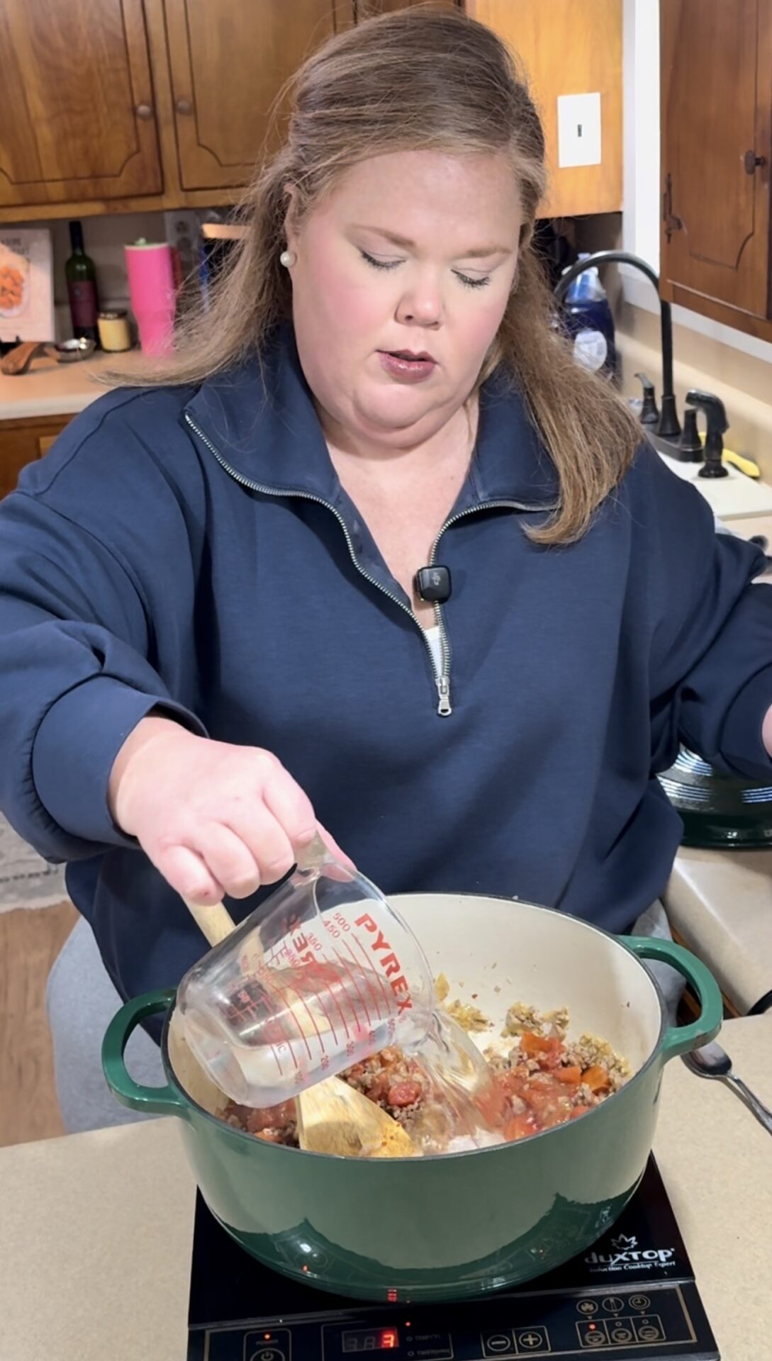 A woman in a navy blue top pours liquid from a measuring cup into a green Dutch oven on an induction stove in a kitchen. Chopped tomatoes and seasoning are visible in the pot.
