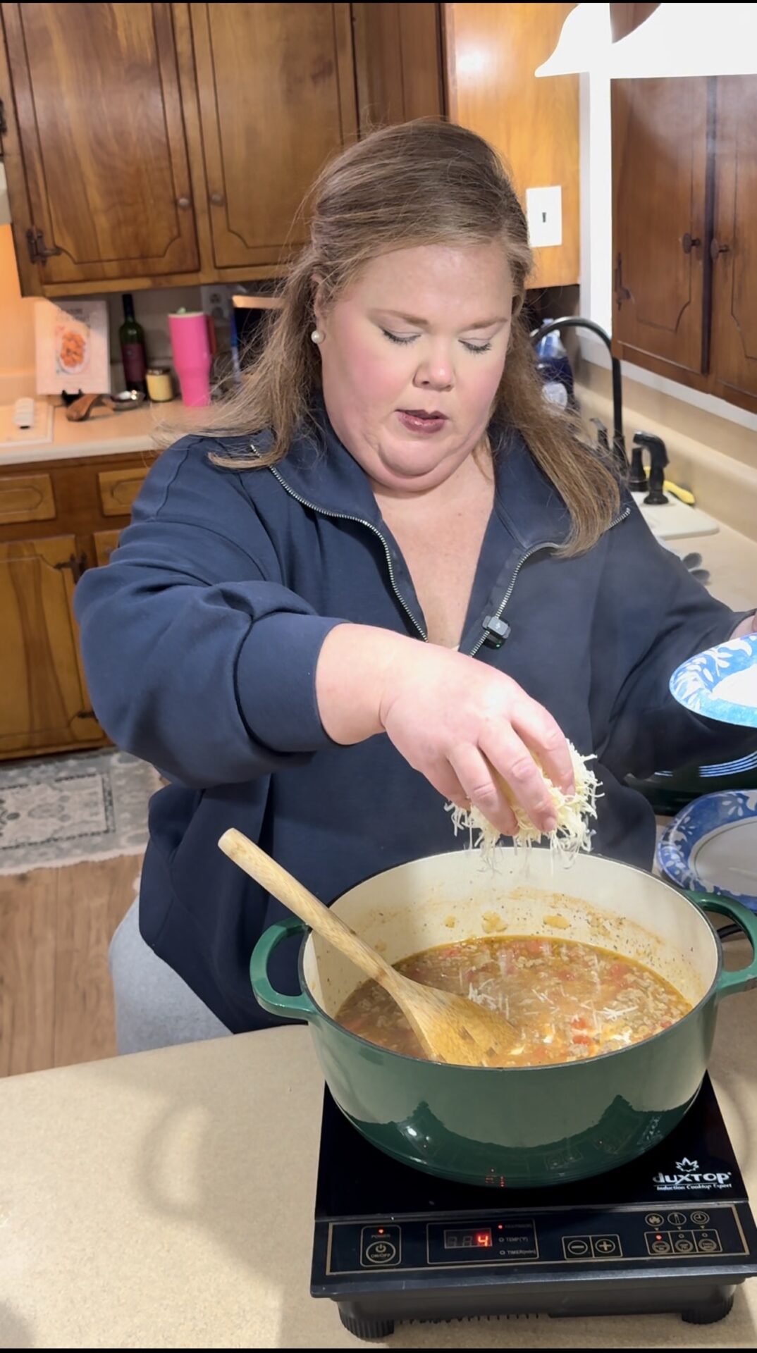 A woman in a navy sweatshirt stands in a kitchen, sprinkling shredded cheese into a pot of soup on the stove. Steam rises from the pot, and a wooden spoon rests inside. Wooden cabinets and kitchen items are visible in the background.