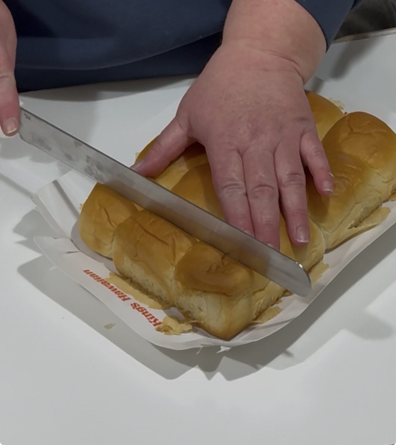 A person uses a large knife to slice a pack of six bread rolls on a white surface, holding the rolls steady with their other hand. Recipe for Hawaiian Roll Grilled Cheese