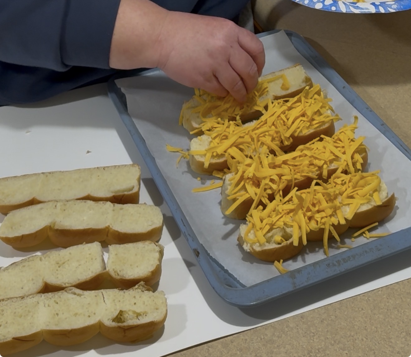 A person sprinkles shredded cheese onto split hoagie rolls arranged on a baking tray lined with parchment paper, preparing the sandwiches for baking.