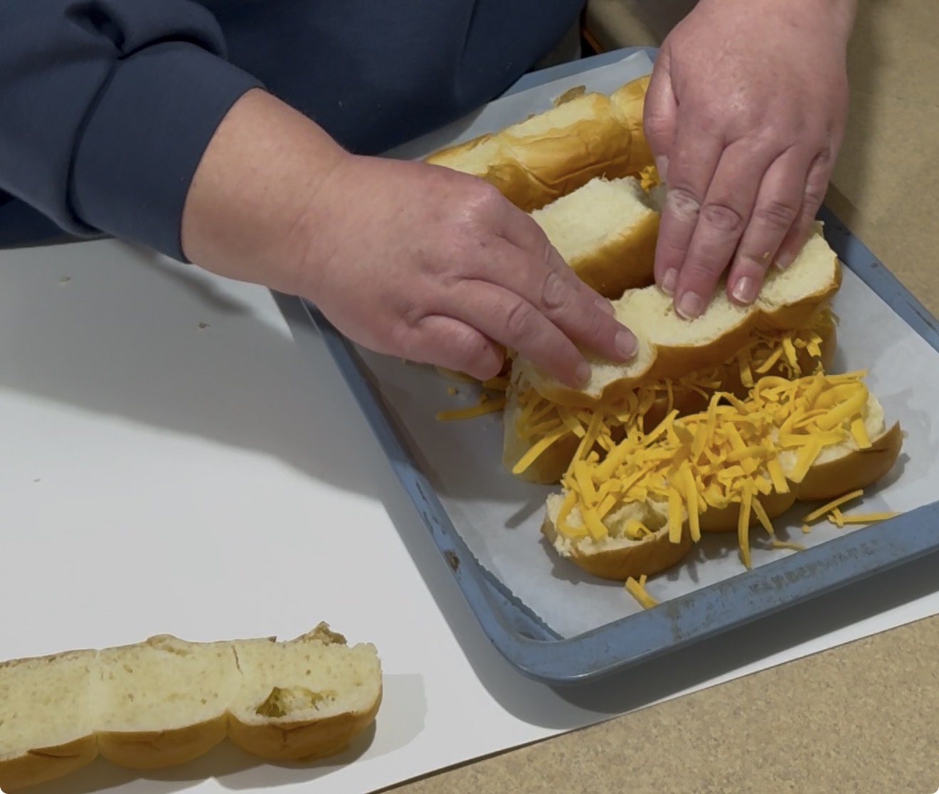 A person places the top half of a sandwich bun onto shredded cheddar cheese and other ingredients on a baking tray lined with parchment paper. Another sandwich bun lies on the counter nearby.