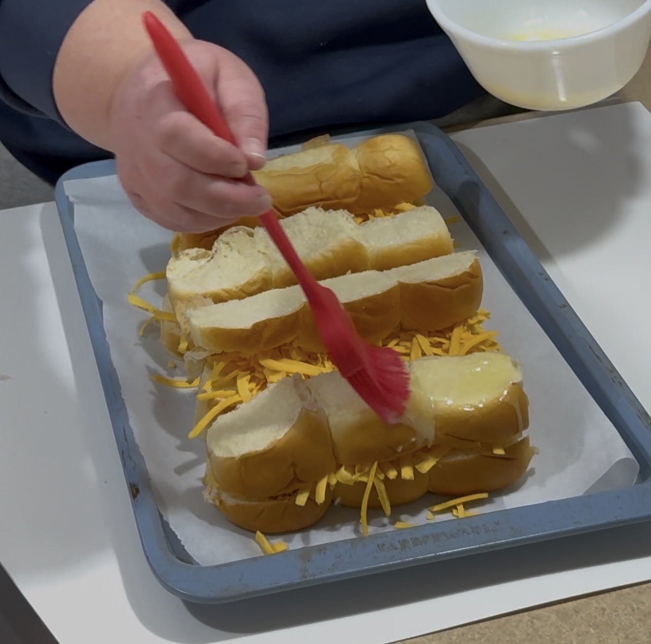 A person brushes melted butter onto sliced hot dog buns filled with shredded cheese on a parchment-lined baking tray.
