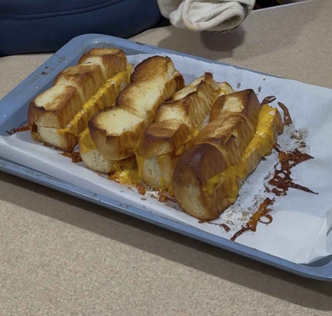 Four grilled cheese sandwiches made with thick slices of bread and melted cheddar cheese are arranged on a parchment-lined baking tray on a countertop. A persons gloved hand and sleeve are visible in the background.