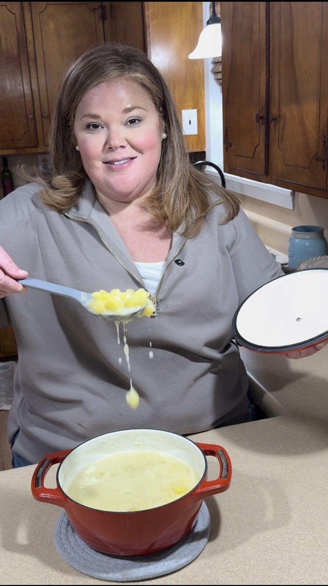 A woman in a gray shirt stands in a kitchen, smiling while holding the lid of a red pot and scooping out a spoonful of soup or stew with potatoes. The pot sits on the countertop, and cabinets are visible in the background.