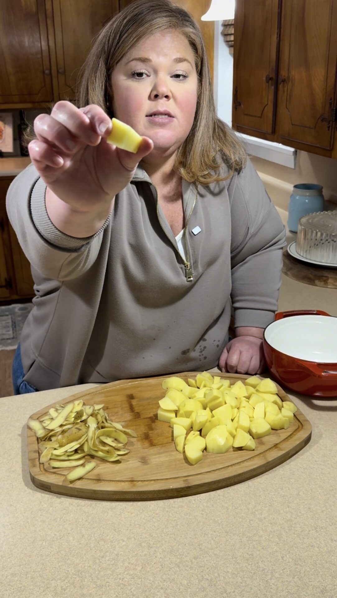 A woman in a kitchen holds up a chunk of peeled potato toward the camera. On the counter in front of her is a cutting board with peeled, chopped potatoes, potato peels, and a red pot.