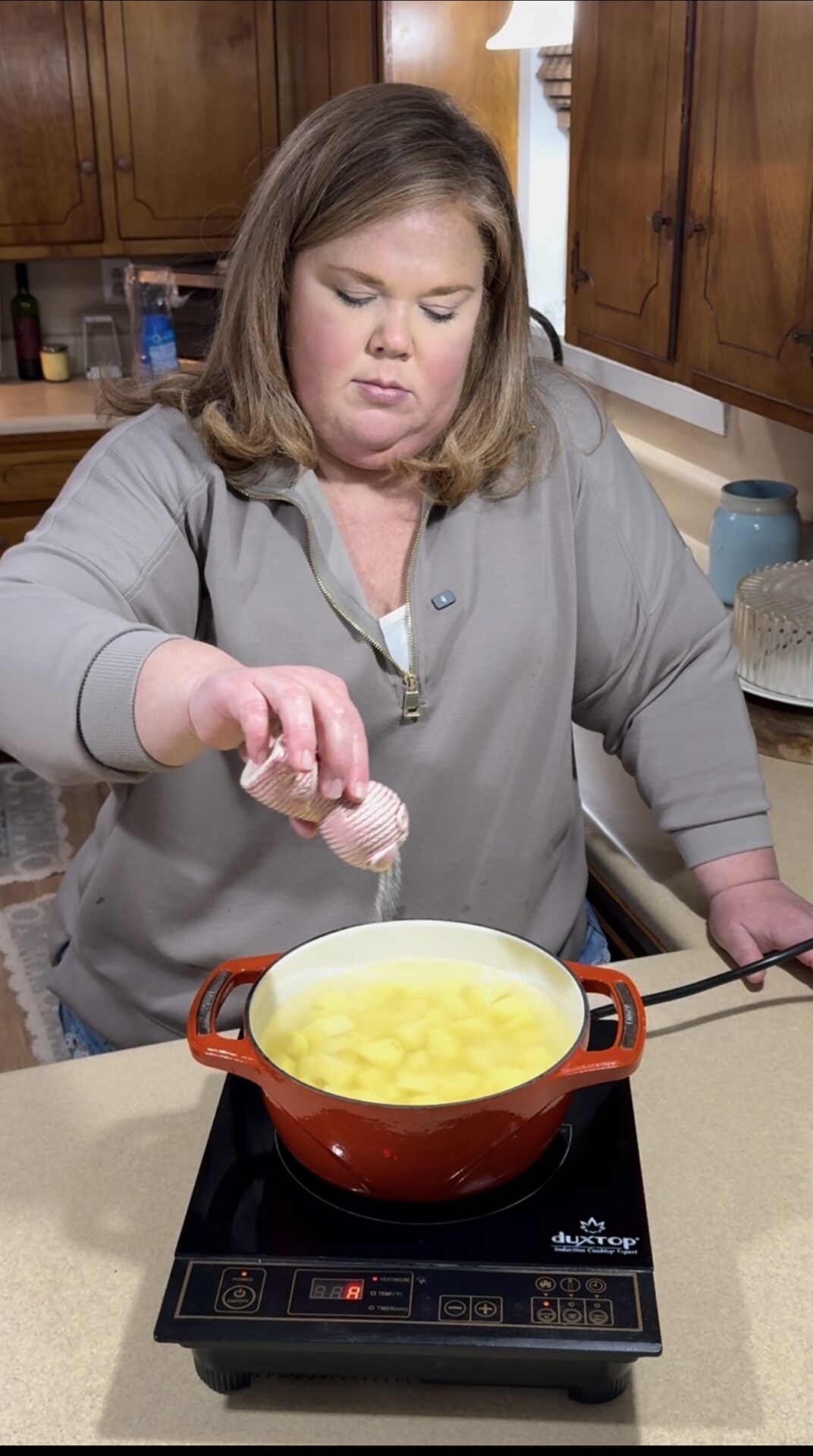 A woman sprinkles salt into a red pot of cubed potatoes boiling on an electric stovetop in a kitchen with wooden cabinets.