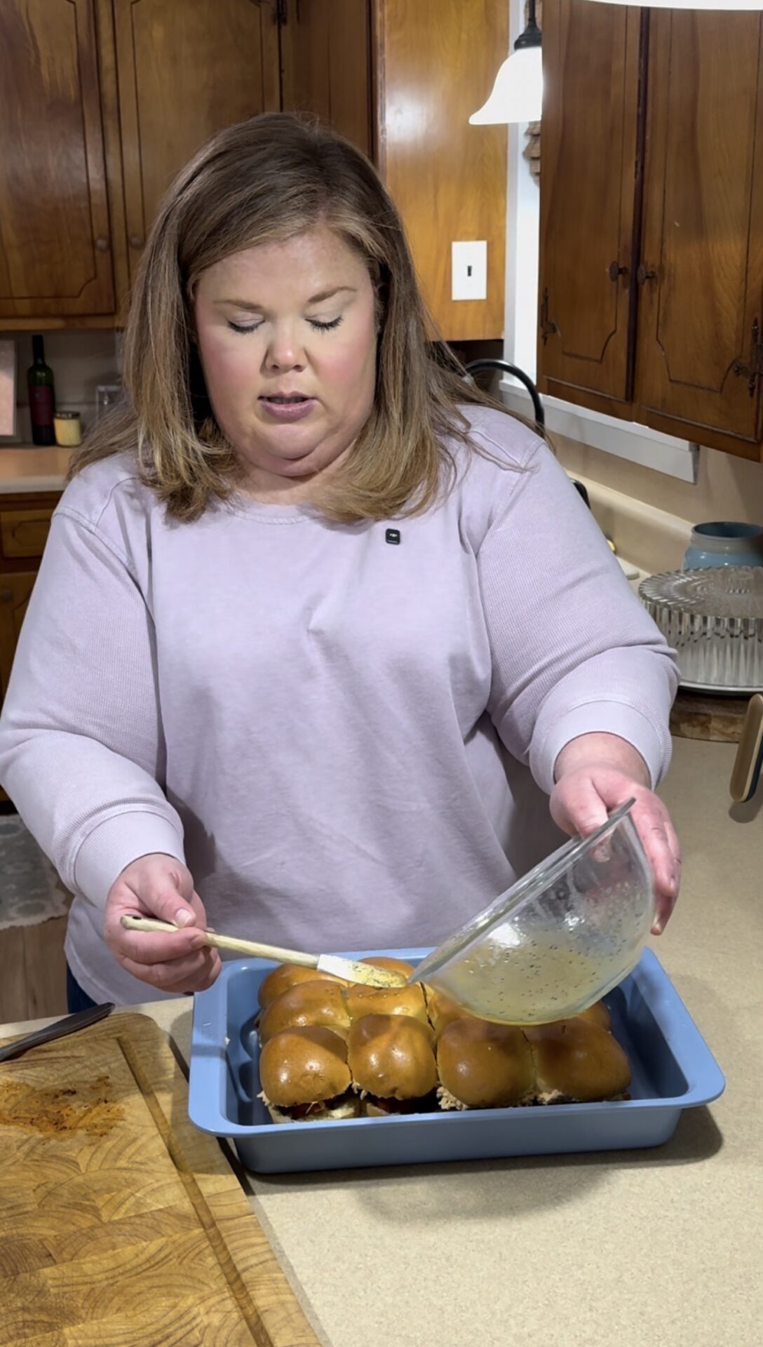 A woman in a kitchen is pouring a liquid mixture from a glass bowl onto a tray of slider sandwiches with shiny buns. Wooden cabinets and kitchen items are visible in the background.
