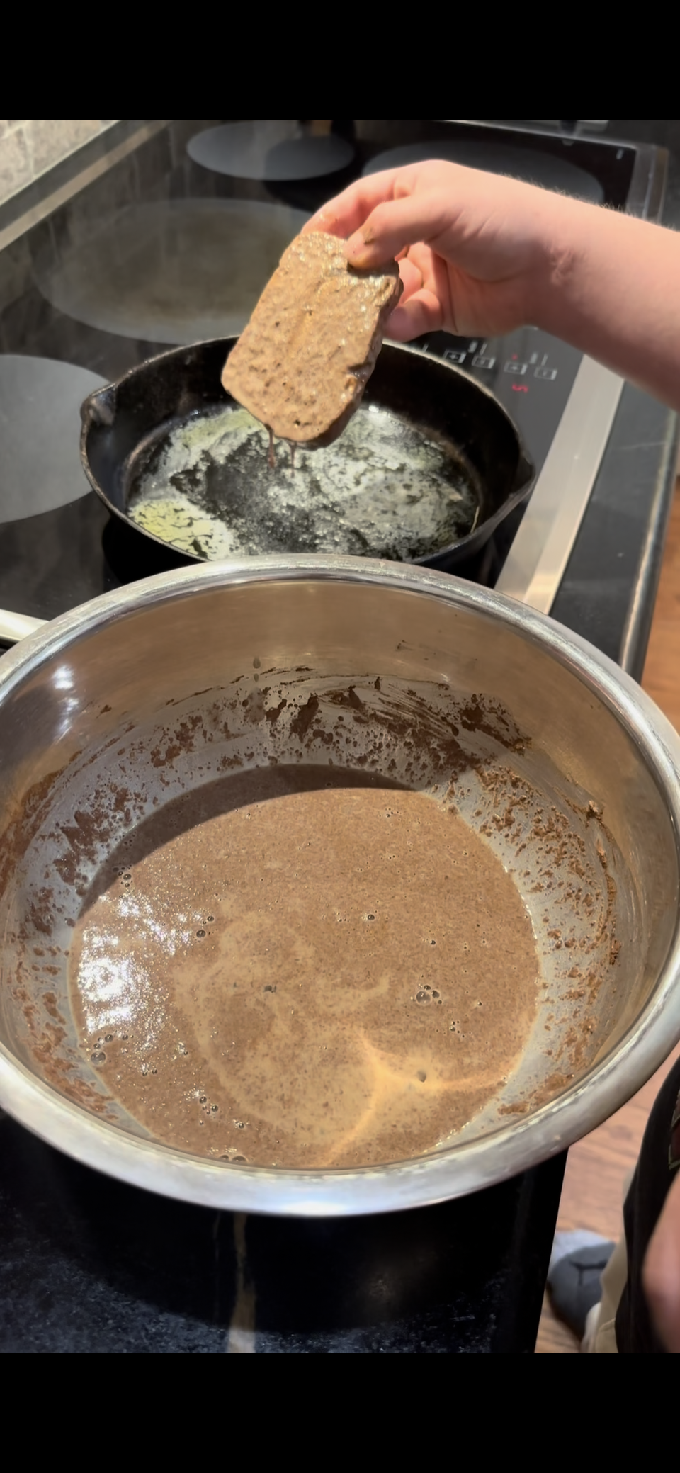 A hand dips a slice of bread into a bowl of brown mixture, preparing to cook it in a skillet of melted butter on a stovetop.
