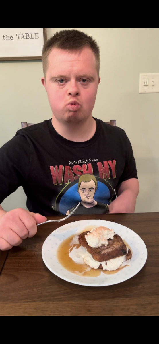 A young man sits at a table with pancakes topped with whipped cream and syrup. He holds a fork and wears a black graphic T-shirt, looking straight at the camera with a slight pout.