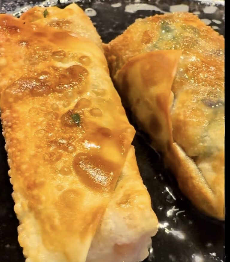 Two golden-brown, crispy egg rolls are frying in oil, with blistered surfaces and visible bubbling, on a dark skillet.
