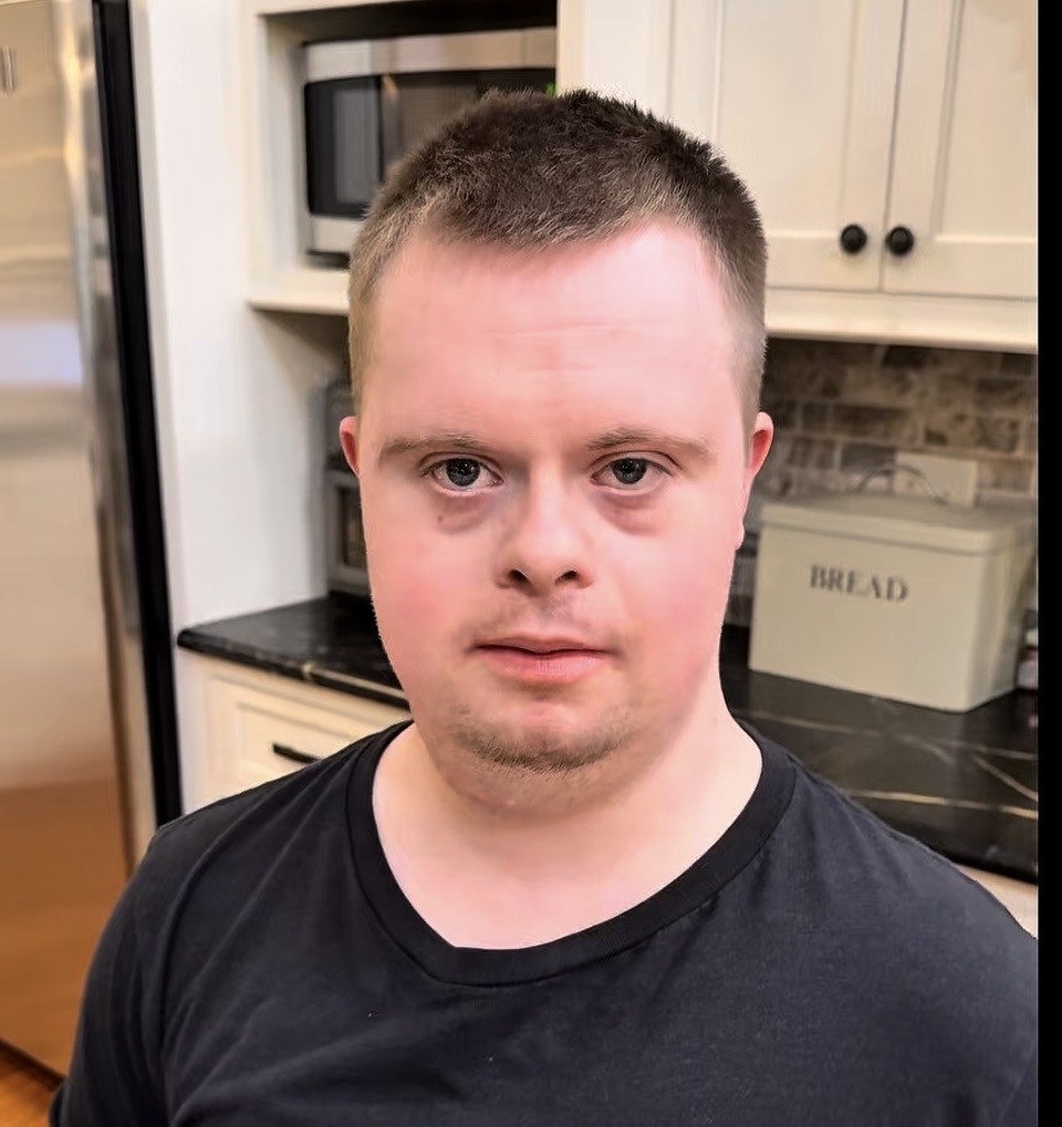A young man with short hair and a black T-shirt stands in a kitchen, looking at the camera. Behind him are white cabinets, a bread box, a microwave, and a stainless steel refrigerator.