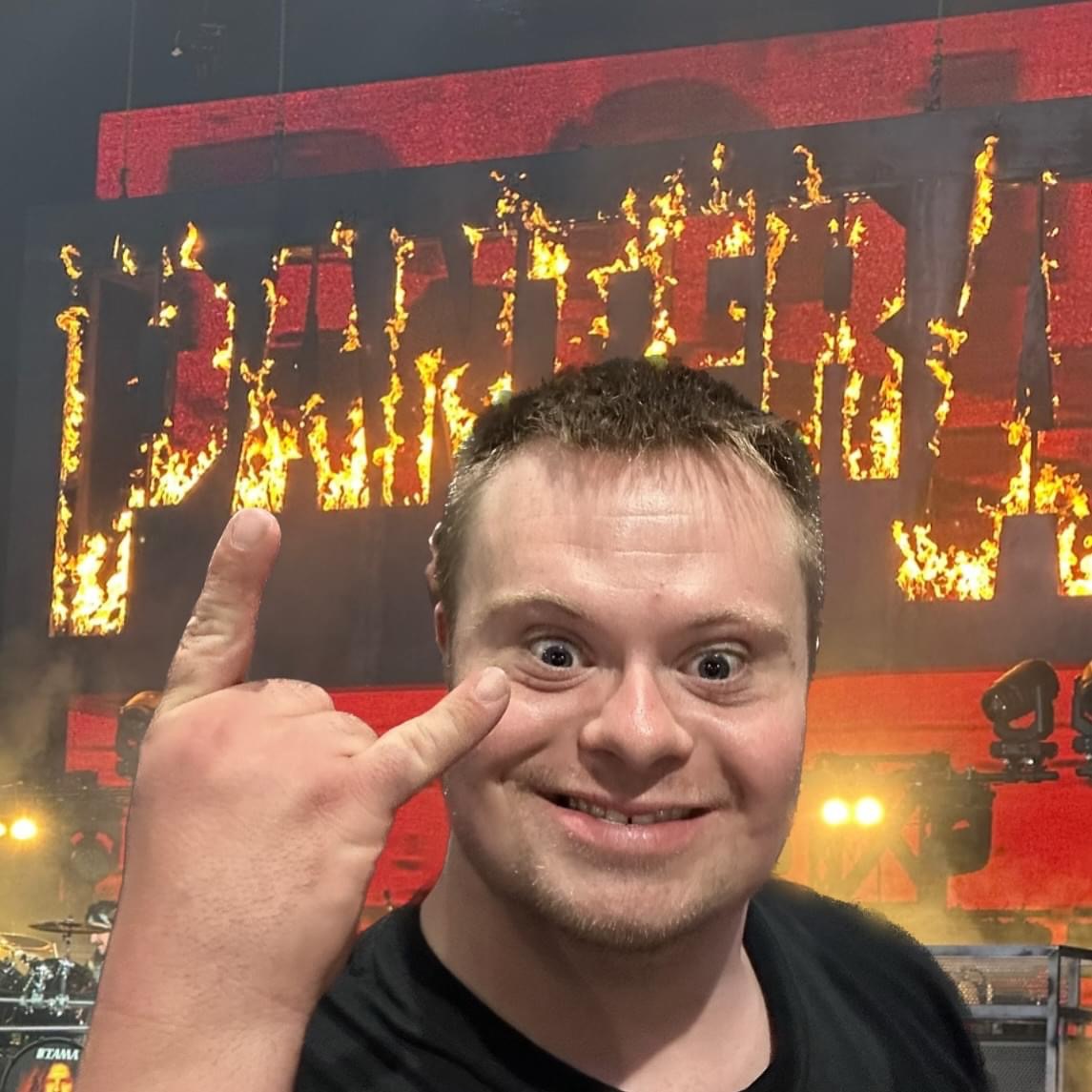 A smiling man makes a rock on hand gesture at a concert, with the word PANTERA displayed in burning letters on a large screen behind him. Bright stage lights and equipment are visible in the background.