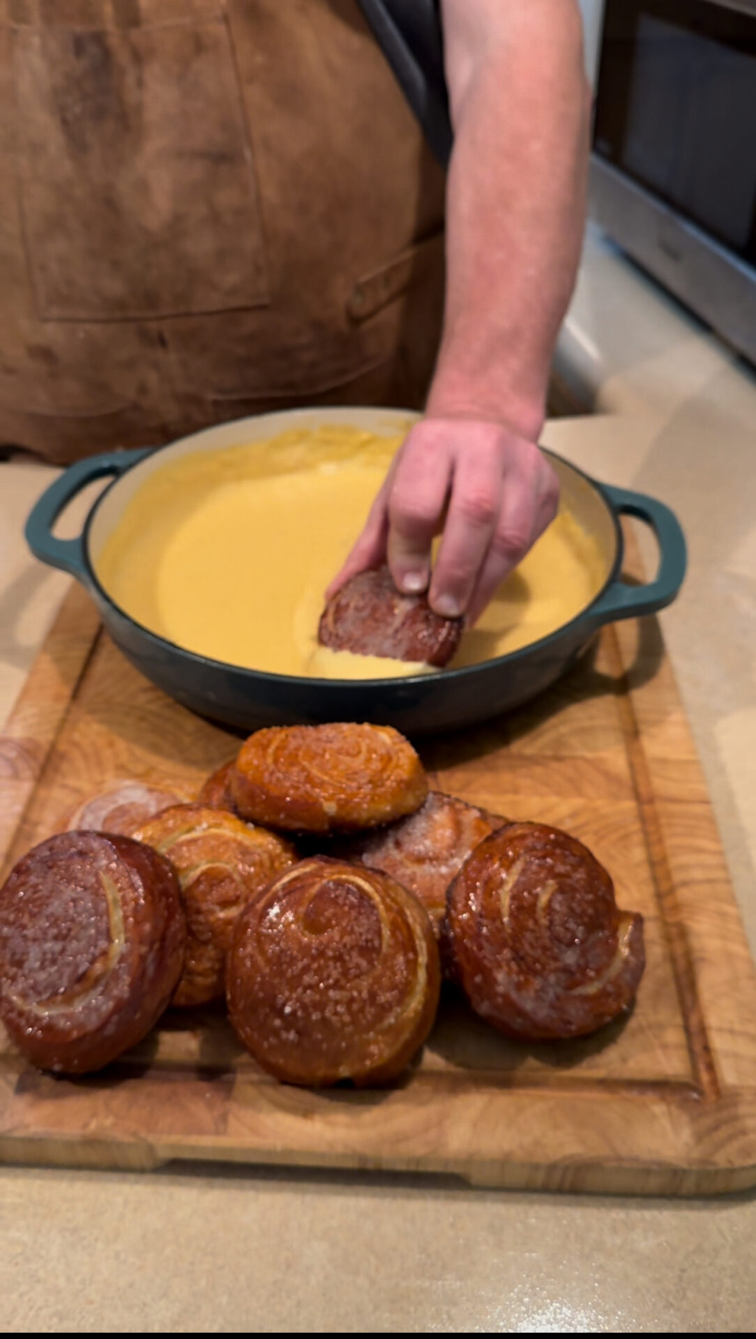 A person dips a round pastry into a pot of yellow sauce, with several similar pastries stacked on a wooden cutting board in the foreground. The person is wearing a brown apron.
