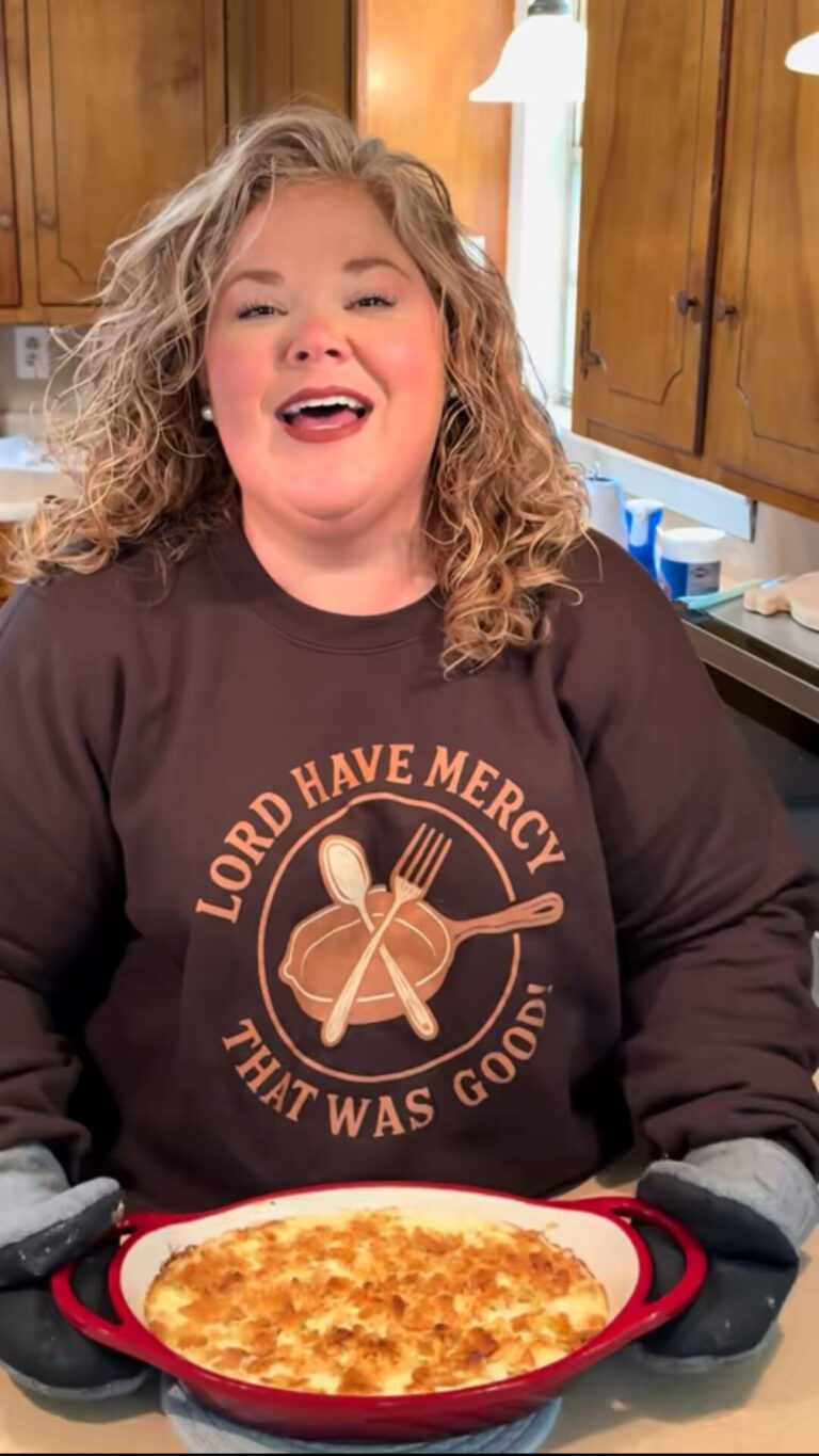 A smiling woman with curly hair holds a red casserole dish of baked food in a kitchen. She wears oven mitts and a sweatshirt that says, LORD HAVE MERCY THAT WAS GOOD! with utensils and food graphics.