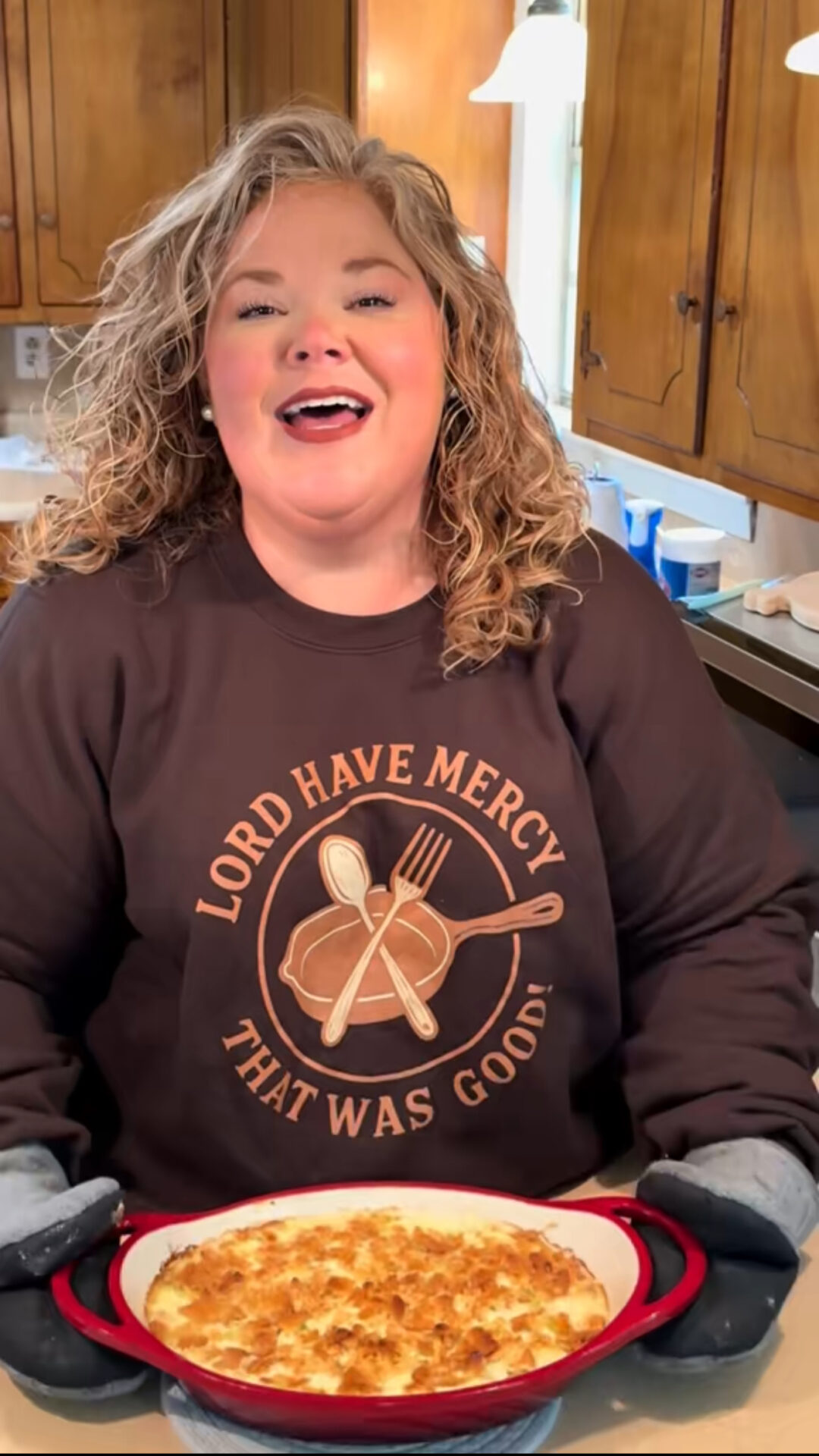 A smiling woman with curly hair holds a red casserole dish of baked food in a kitchen. She wears oven mitts and a sweatshirt that says, LORD HAVE MERCY THAT WAS GOOD! with utensils and food graphics.