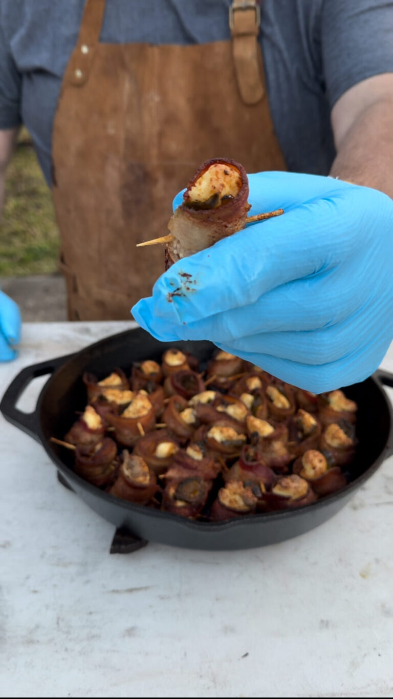 A person wearing blue gloves and a brown apron holds up a bacon-wrapped appetizer on a toothpick over a cast iron skillet filled with similar appetizers.