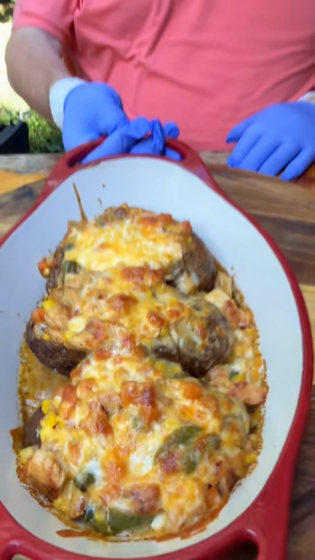 A person wearing a coral shirt and blue gloves holds a red and white baking dish filled with baked stuffed potatoes topped with melted cheese and various vegetables.