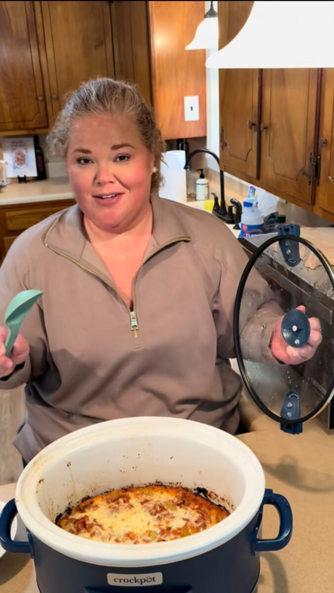 A woman stands in a kitchen holding a spoon and the lid of a blue Crockpot, which contains a baked cheesy dish. She is smiling and wearing a beige pullover. Wooden cabinets and a sink are visible in the background.