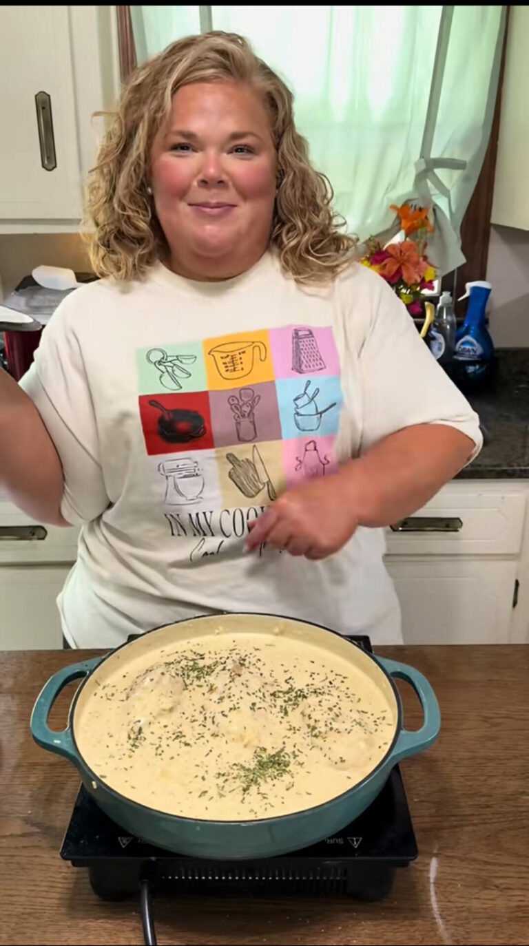 A smiling woman with curly blonde hair stands in a kitchen, pointing at a large pot of creamy pasta topped with herbs on a countertop. She wears a white shirt with colorful kitchen-themed illustrations.