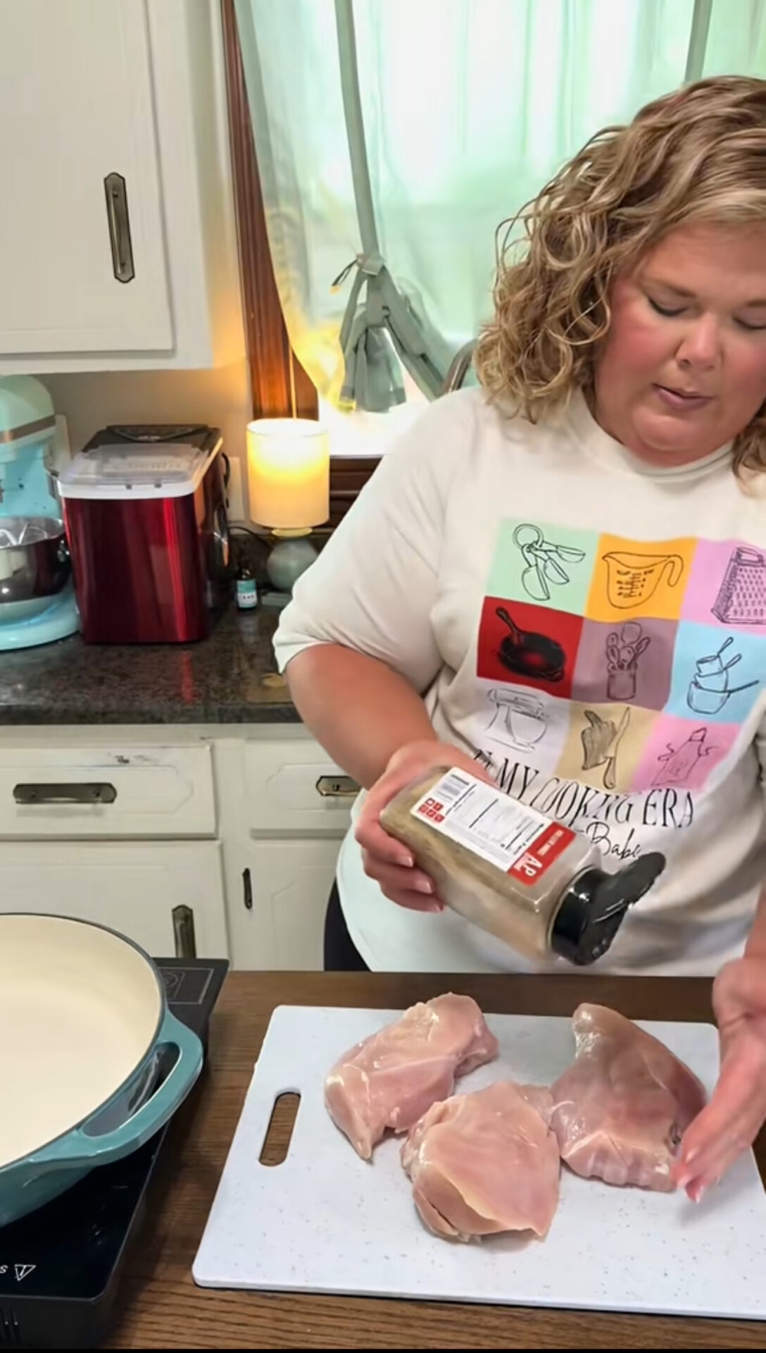 A woman in a colorful graphic shirt seasons raw chicken breasts on a cutting board in a kitchen. A pot sits on the stove beside her, and kitchen appliances are visible in the background.