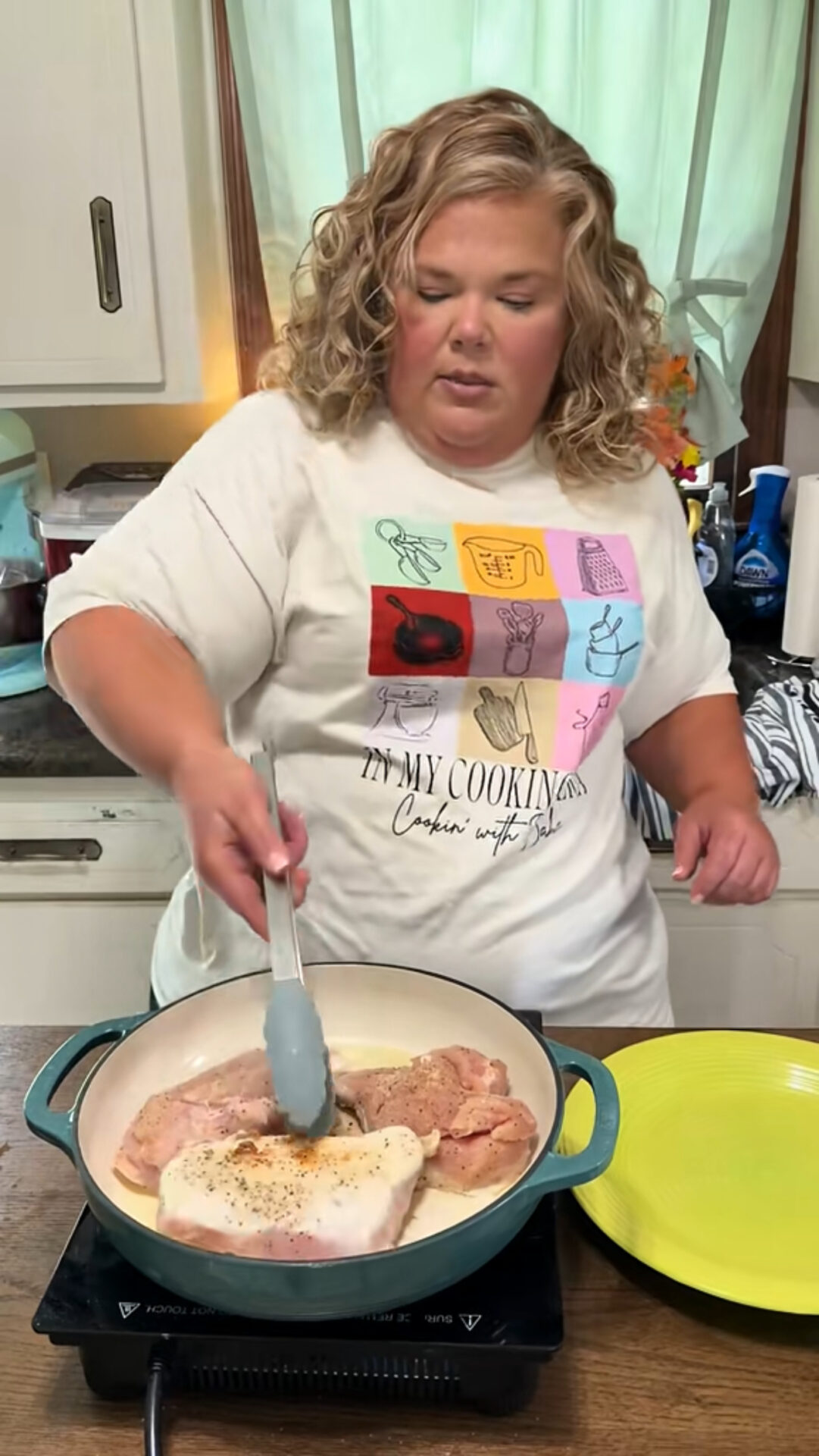 A woman with curly blonde hair, wearing a graphic t-shirt, uses tongs to cook seasoned meat in a skillet on a stovetop in a kitchen. A yellow plate sits beside her, and light streams in from a window behind.