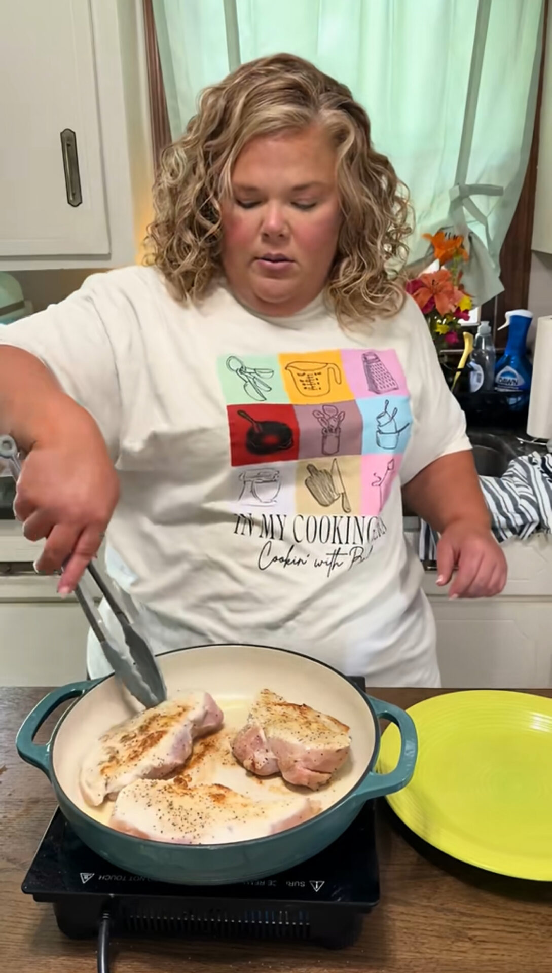 A woman with curly blonde hair cooks seasoned pork chops in a white skillet using tongs, standing in a kitchen. She wears a white graphic T-shirt and is focused on flipping the meat. A yellow plate sits nearby.