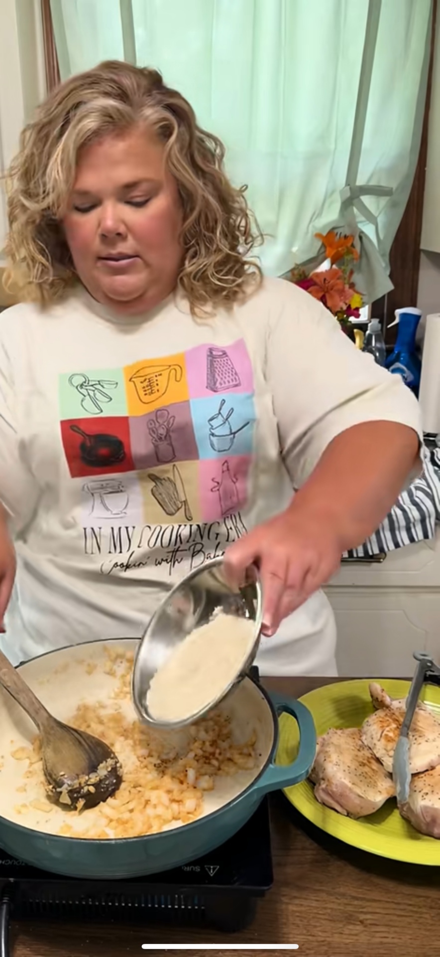 A woman with curly blonde hair cooks in a kitchen, pouring a white powder from a bowl into a pot of sautéed onions on the stove. She wears a white shirt with colorful graphic drawings. Cooked chicken is on a green plate nearby.