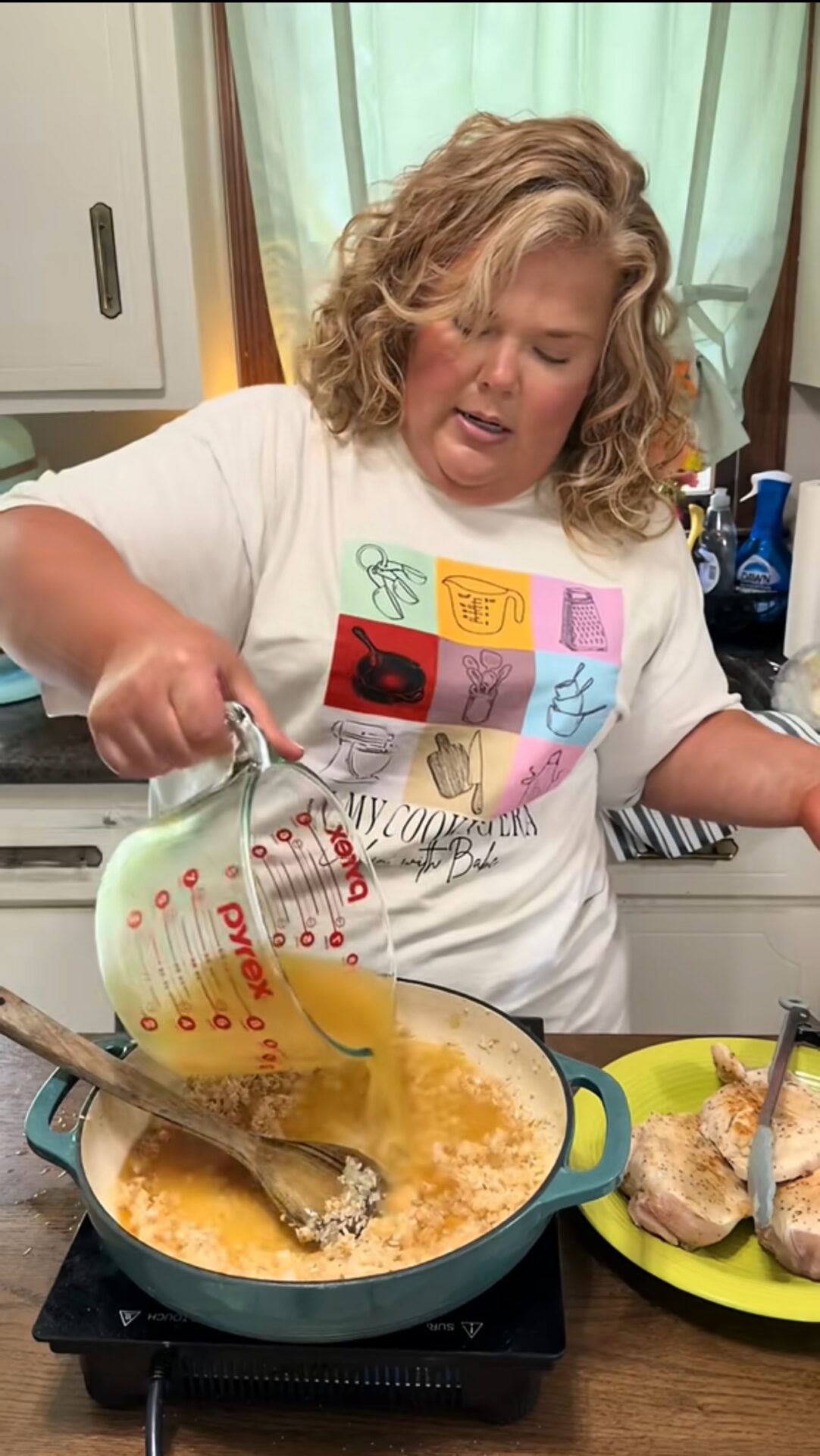 A woman with curly blonde hair pours broth from a Pyrex measuring cup into a pot of rice on the stove. Cooked chicken breasts are on a plate beside her in a kitchen with white cabinets and a window.