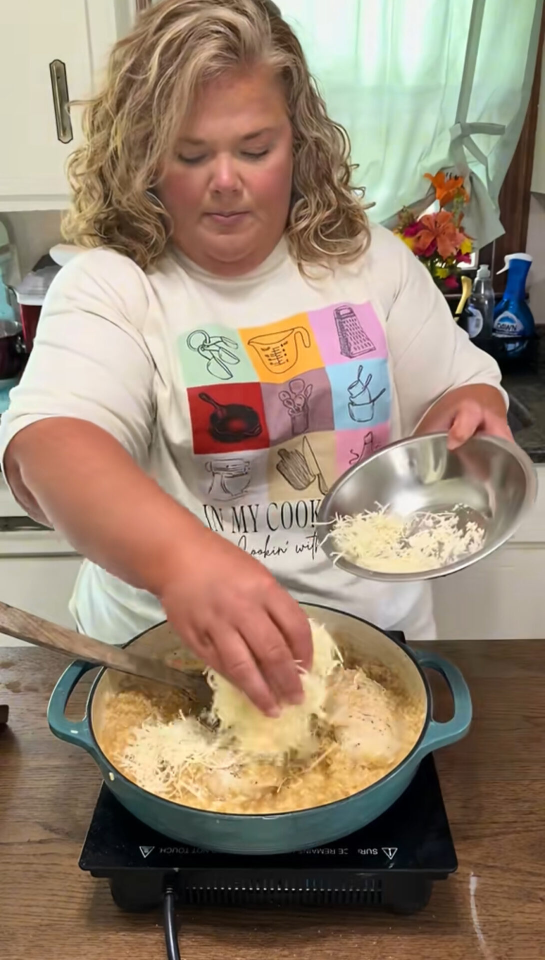 A woman with curly blonde hair adds shredded cheese from a bowl into a large pot on a stovetop in a kitchen. She is wearing a white shirt with colorful kitchen-themed illustrations.