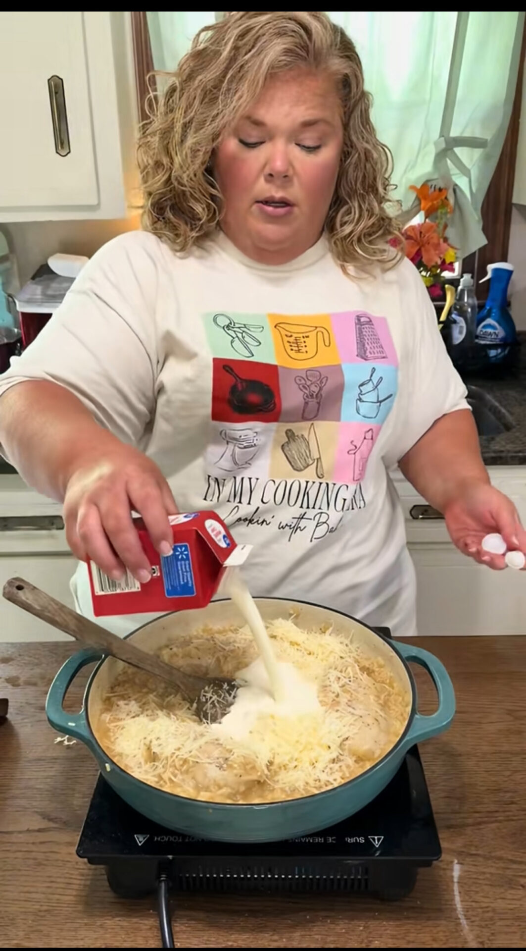 A woman with curly blonde hair pours cream into a pot of food on a stove in a kitchen. She wears a white graphic T-shirt and stands in front of a window with light green curtains.