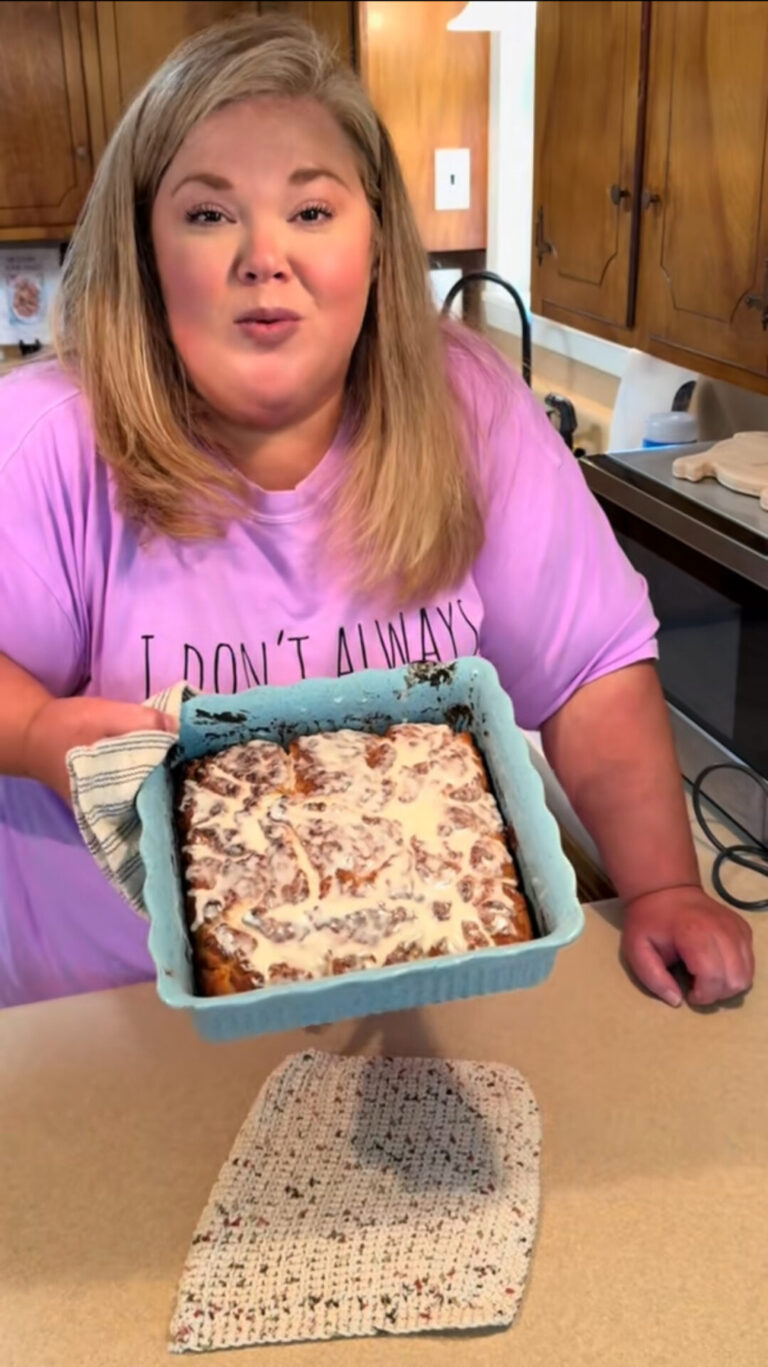 A woman with long blonde hair wearing a lavender shirt holds a blue baking dish filled with iced cinnamon rolls in a kitchen, smiling at the camera.