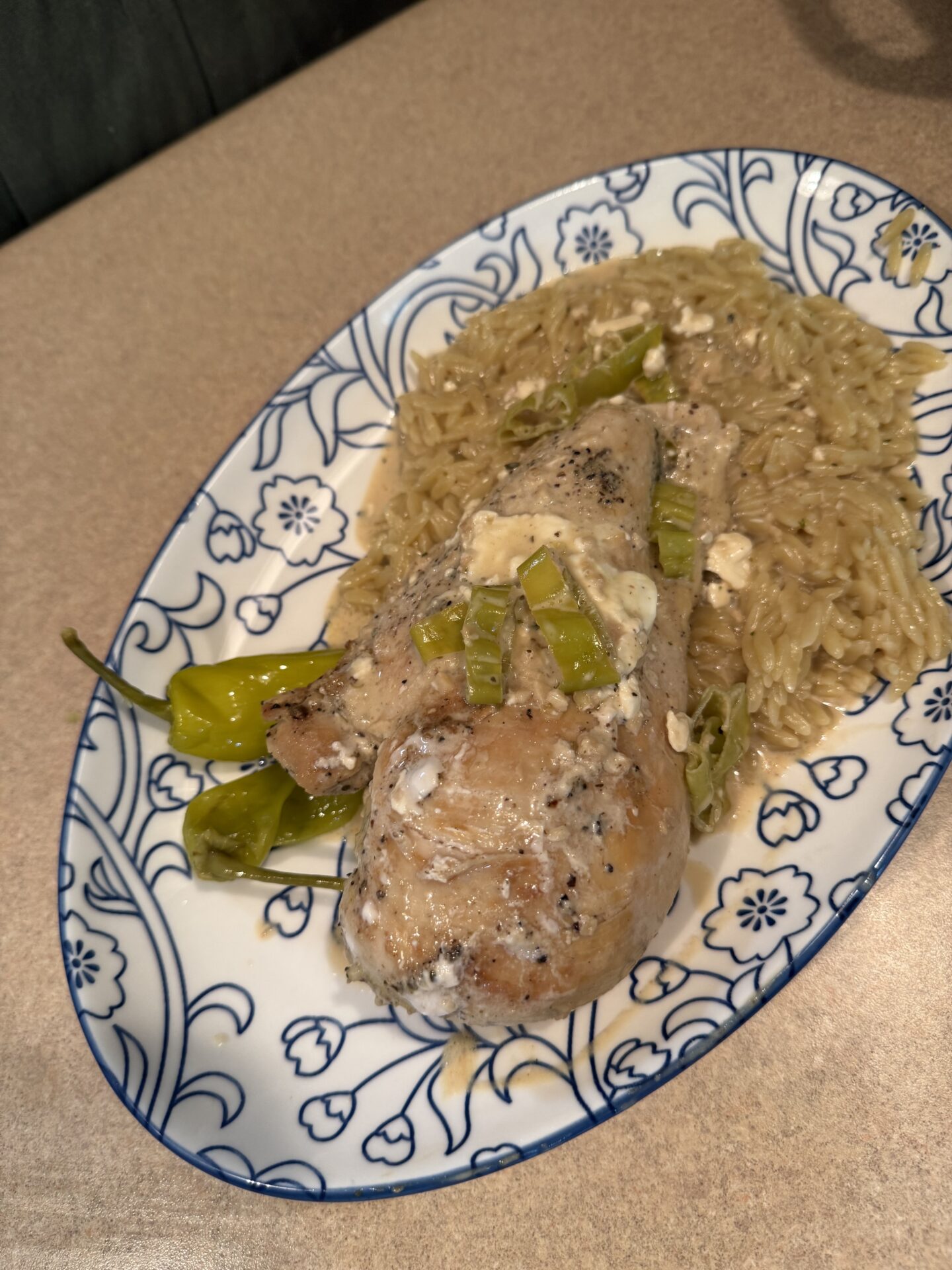 A serving of cooked orzo pasta topped with a seasoned baked chicken leg, sliced green peppers, and a light creamy sauce, presented on a blue and white patterned plate.