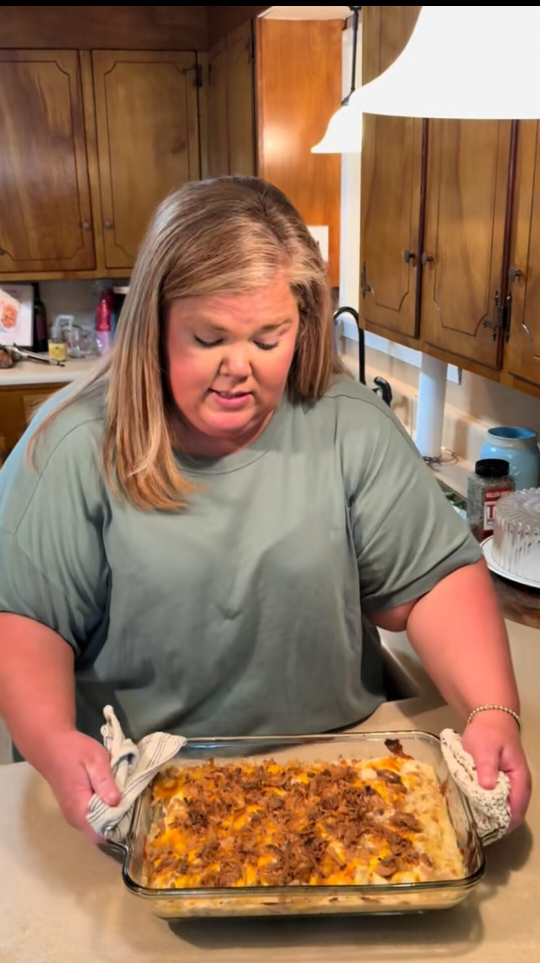 A woman in a light green shirt holds a glass baking dish with a casserole topped with melted cheese and meat in a kitchen with wooden cabinets.