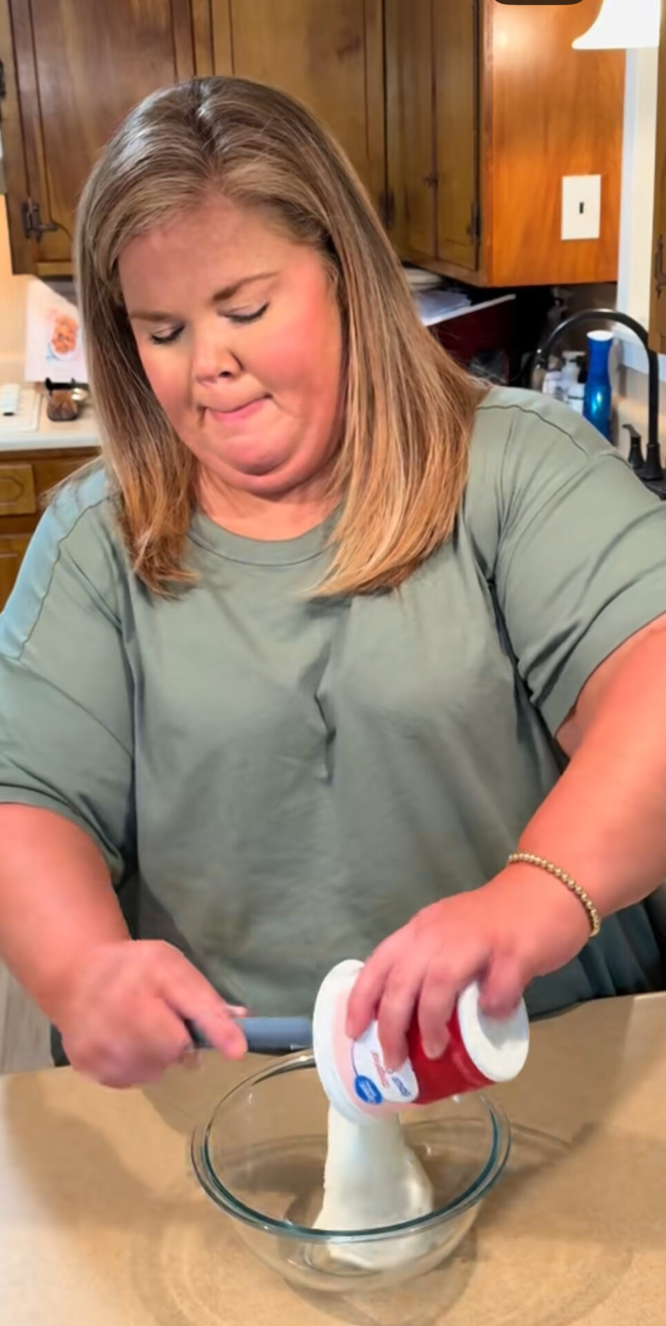 A woman in a green shirt stands in a kitchen, spooning contents from a white and red container into a clear glass bowl on the counter.