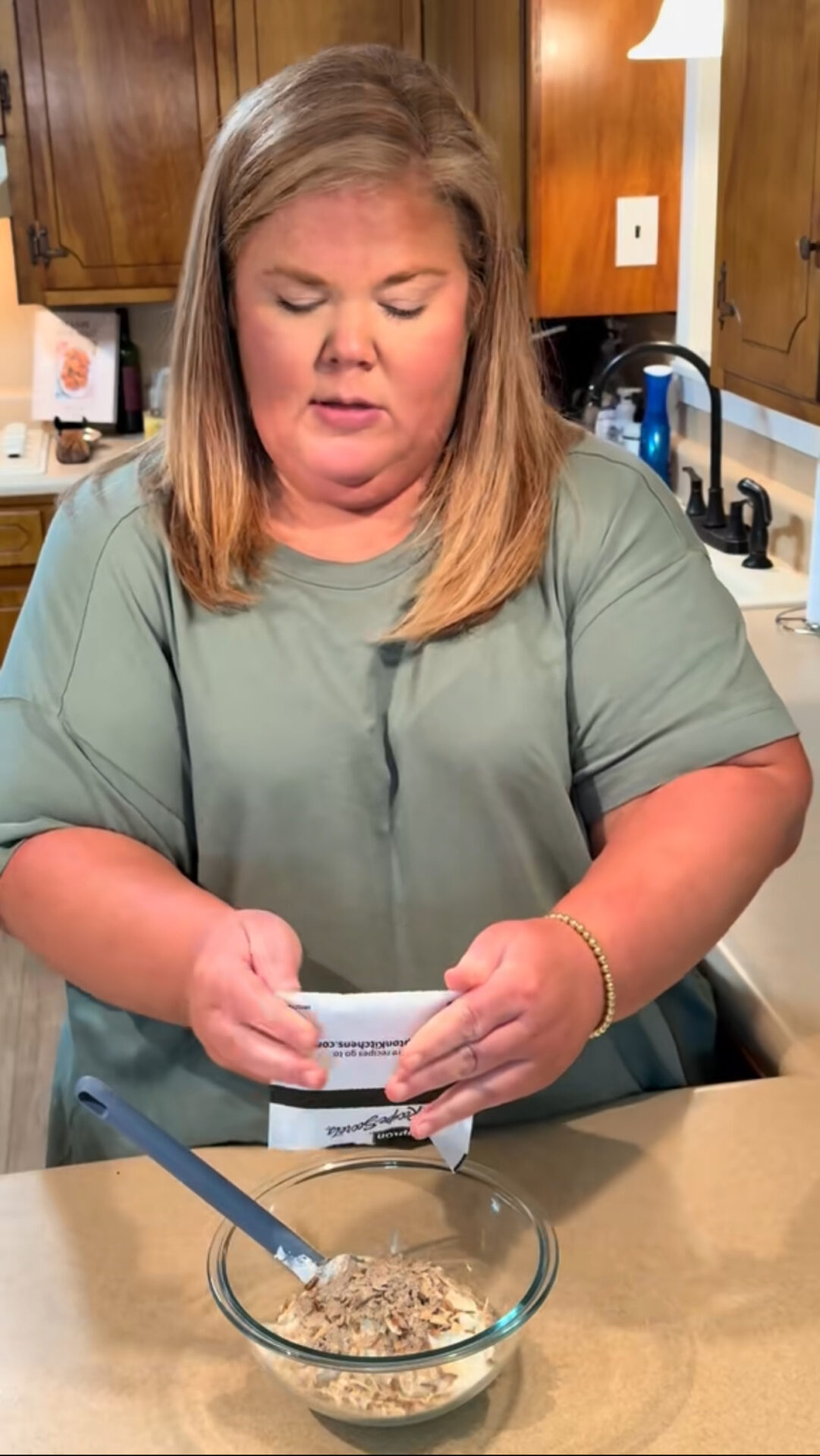 A woman stands in a kitchen pouring the contents of a packet into a glass bowl filled with dry ingredients. A spoon rests in the bowl, and wooden cabinets are visible in the background.