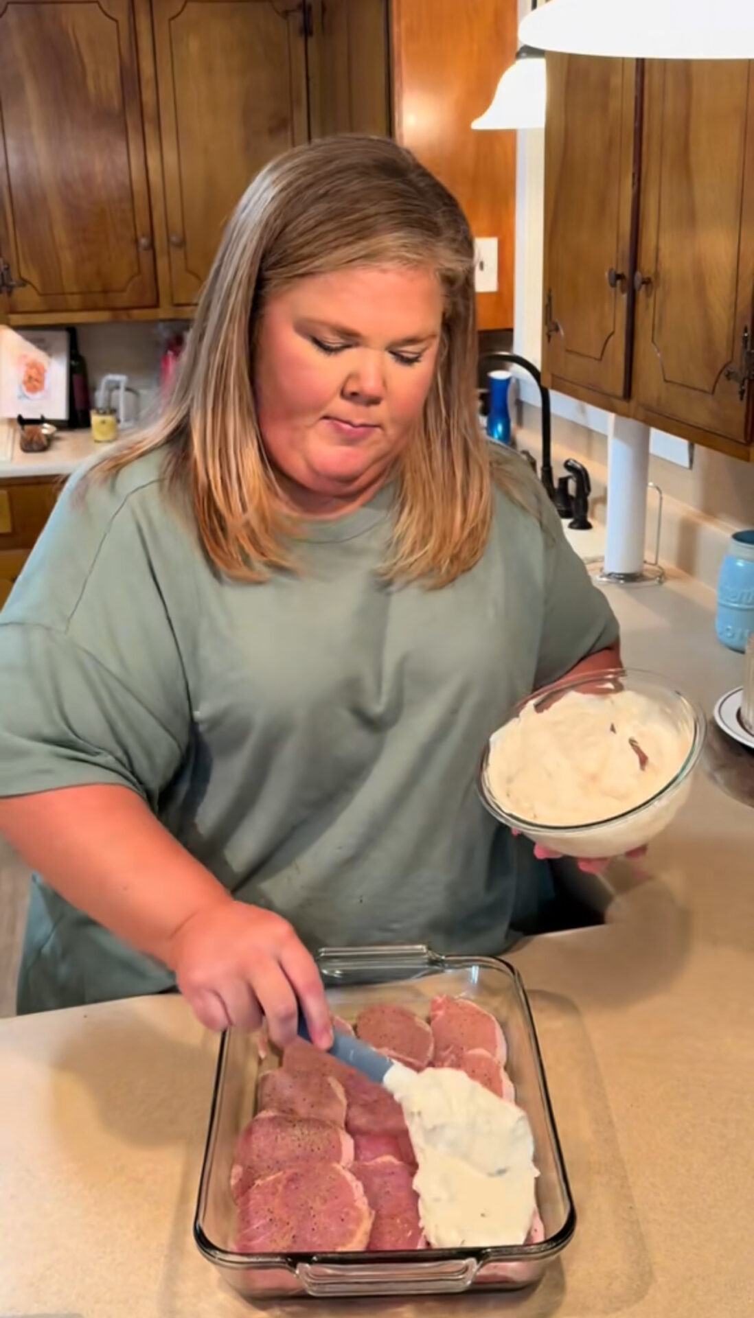 A woman in a green shirt spreads a creamy white mixture over raw pork chops arranged in a glass baking dish on a kitchen counter. Wooden cabinets and kitchen items are visible in the background.