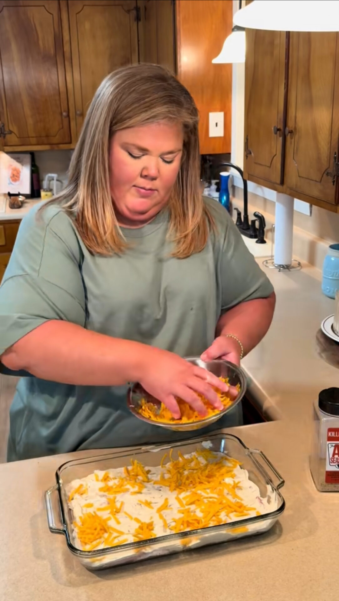 A woman in a green shirt sprinkles shredded cheese from a bowl onto a casserole dish filled with a white mixture in a kitchen with wooden cabinets.