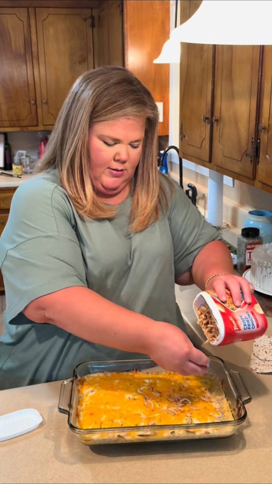 A woman in a green shirt sprinkles French fried onions from a container onto a casserole topped with melted cheese in a glass baking dish in a kitchen.