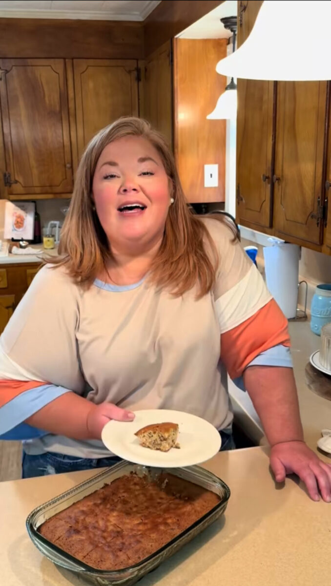 A woman in a kitchen smiles while holding a plate with a slice of cake. A glass baking dish with the remaining cake sits on the counter in front of her. Wooden cabinets and kitchen items are visible in the background.