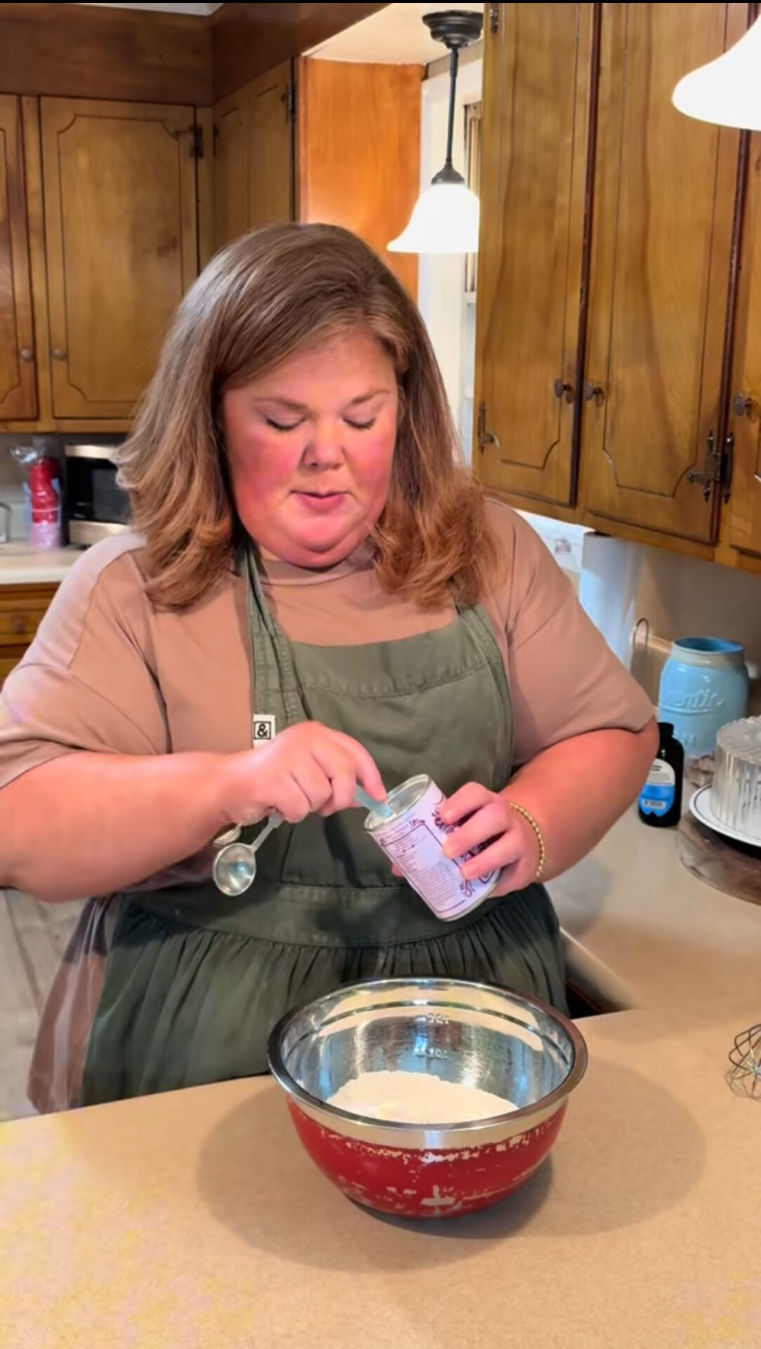 A woman wearing a green apron stands in a kitchen, scooping baking powder from a container into a red mixing bowl on the counter. Wooden cabinets and kitchen items are visible in the background.