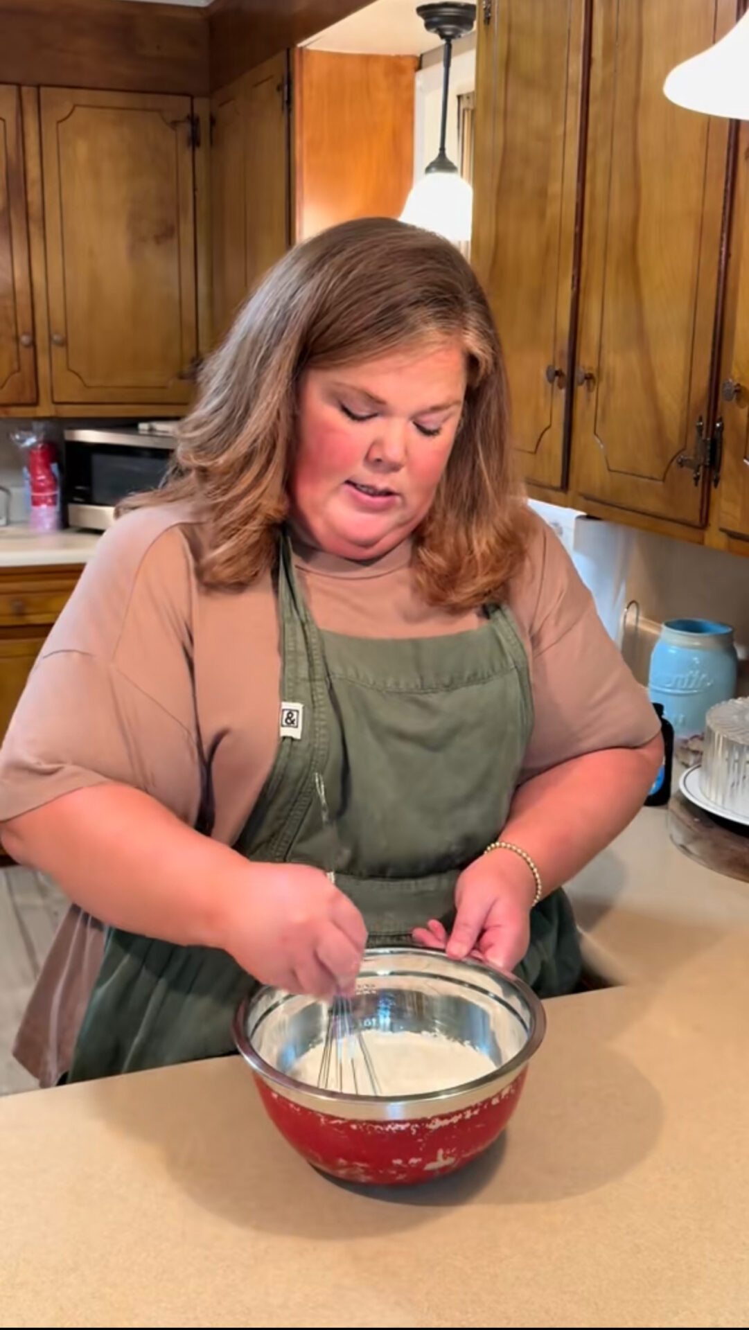 A woman wearing a green apron and brown shirt is whisking ingredients in a red mixing bowl on a kitchen counter, with wooden cabinets and kitchen appliances in the background.
