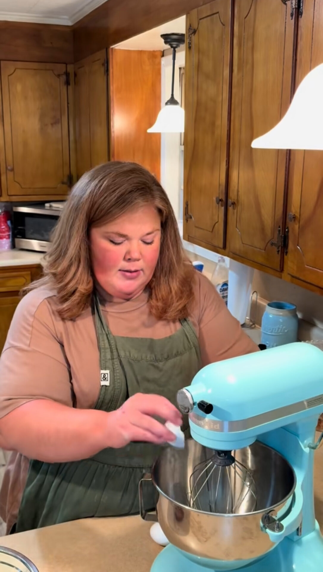 A woman wearing a green apron cracks an egg into a blue stand mixer in a kitchen with wooden cabinets and warm lighting.