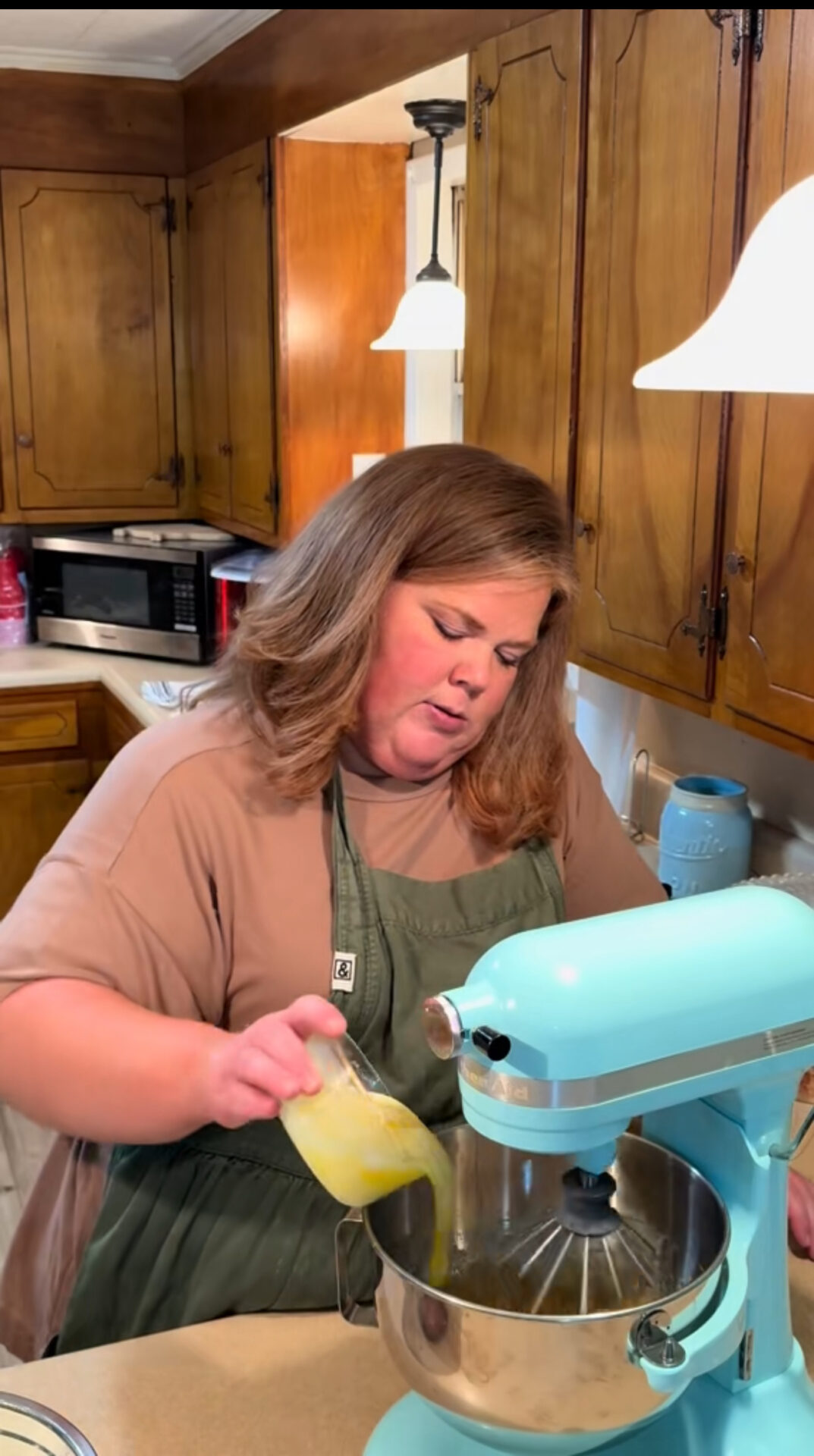 A woman in a green apron pours a yellow liquid into a blue stand mixer in a kitchen with wooden cabinets and hanging lights.