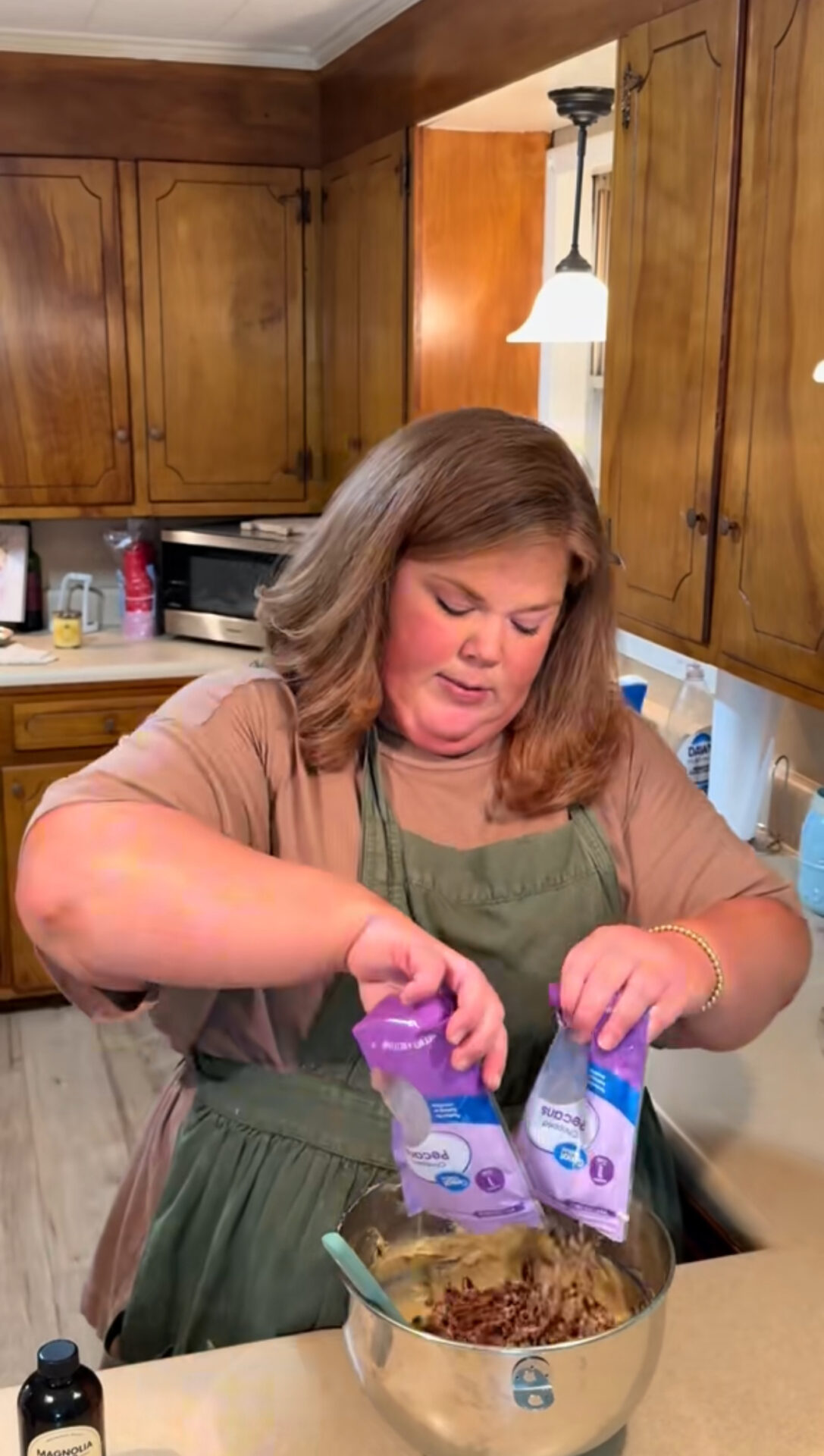A woman in an olive apron stands in a kitchen, pouring two bags of pecans into a large mixing bowl on the counter, preparing ingredients for baking. Brown wooden cabinets and kitchen items are visible in the background.