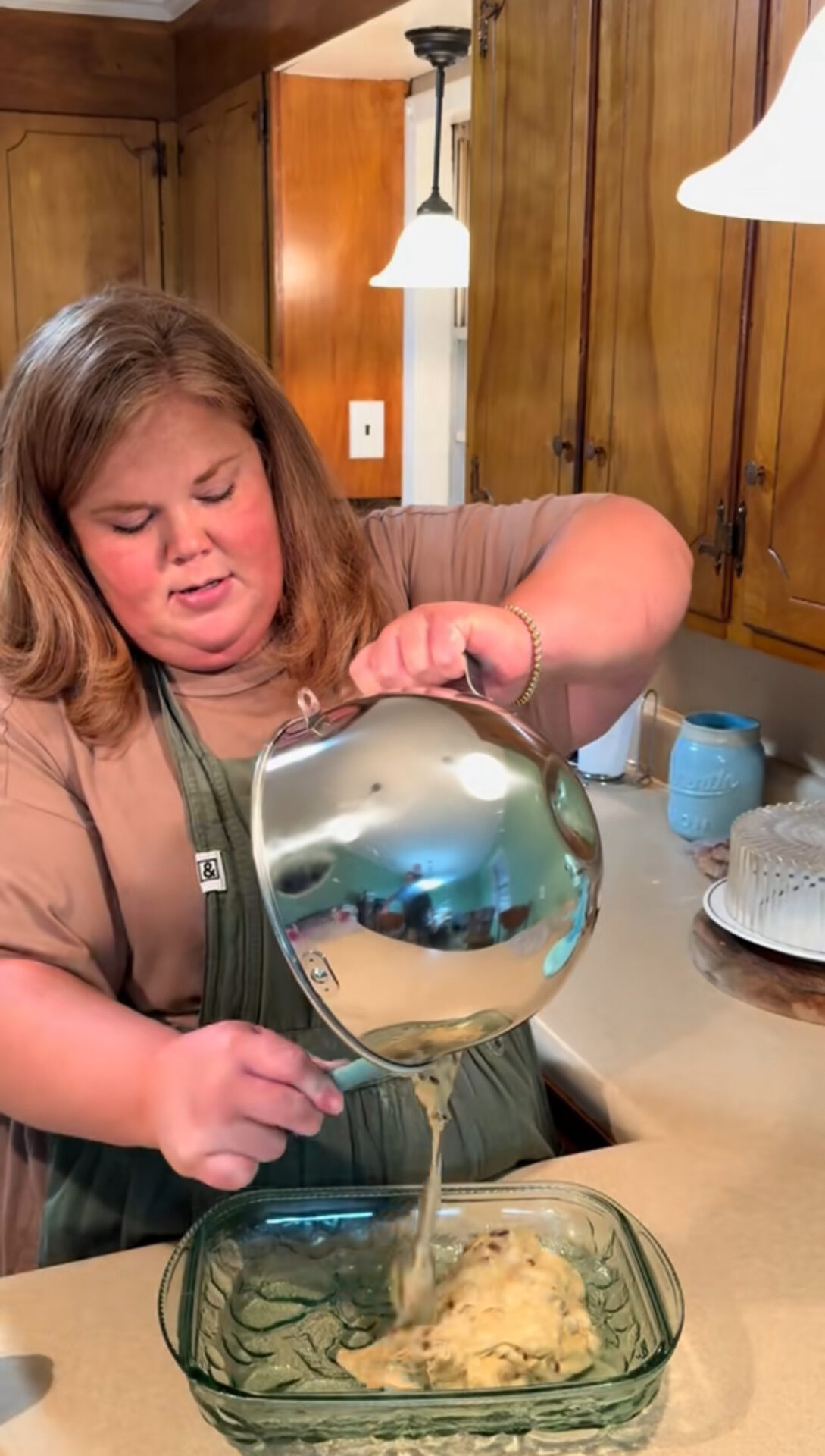 A woman in a kitchen pours batter from a metal mixing bowl into a glass baking dish on the counter, with wooden cabinets and a cake on a stand visible in the background.
