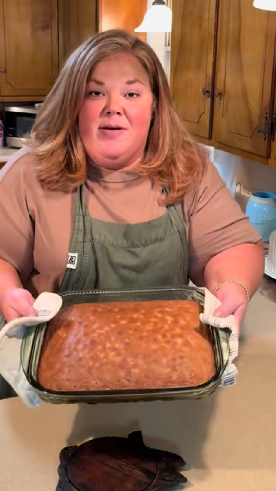 A woman wearing a green apron holds a glass baking dish with a freshly baked cake, standing in a kitchen with wooden cabinets and beige countertops.