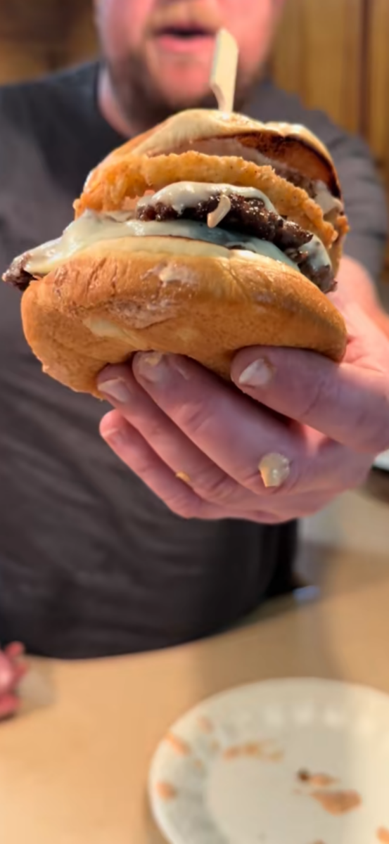 A person holding a large cheeseburger with melted cheese and onion rings, with a bit of sauce on their fingers. The burger is held close to the camera, and a plate is visible on the counter in the background.