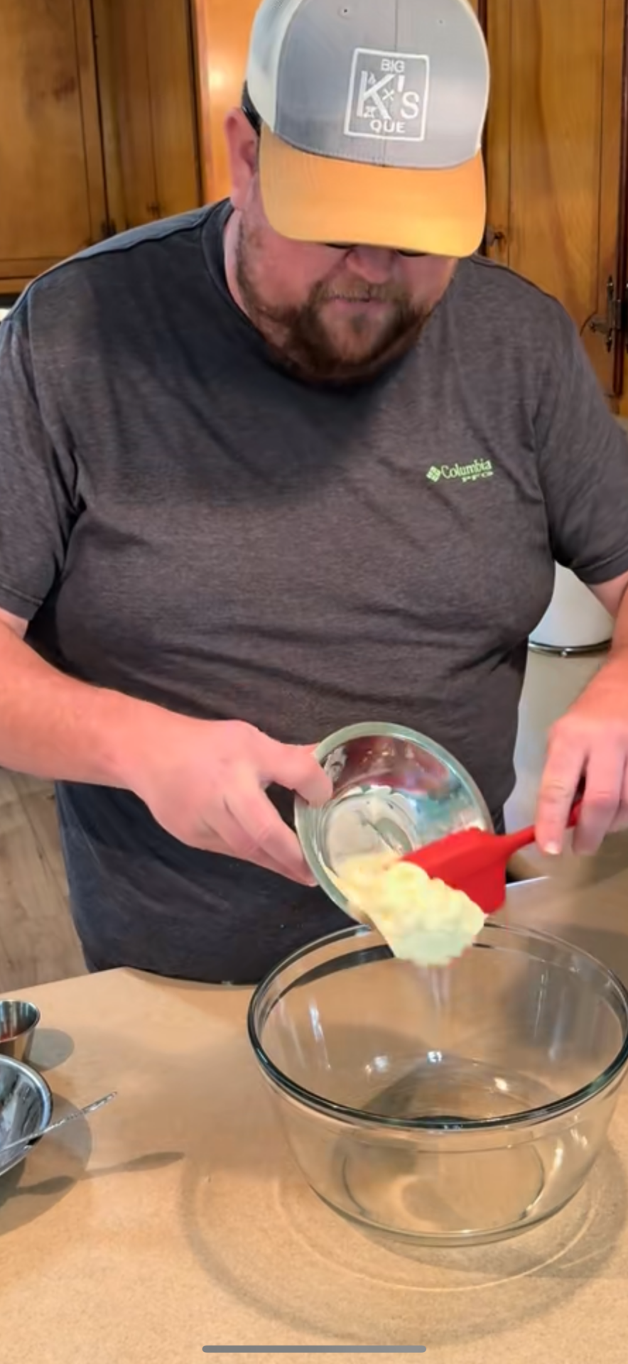 A man wearing a gray hat and shirt pours a thick, creamy mixture from a glass bowl into a large clear mixing bowl on a kitchen counter.