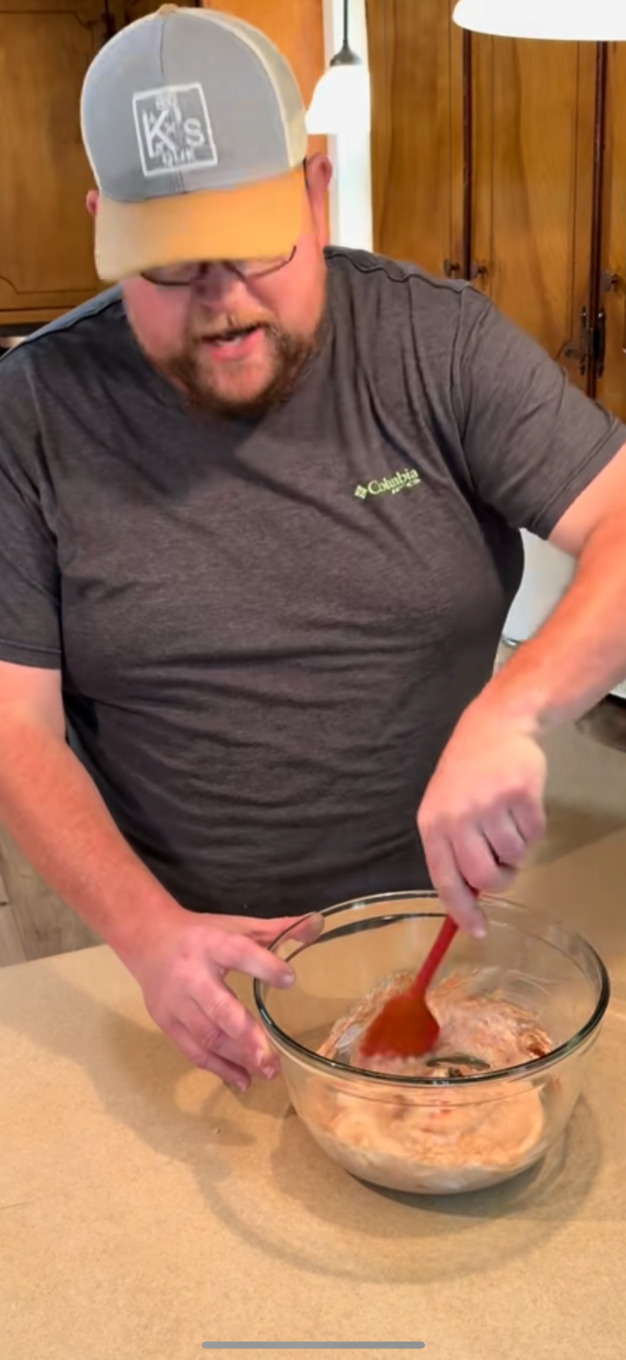 A man wearing glasses, a gray t-shirt, and a cap stirs ingredients in a glass bowl with a red spatula on a kitchen counter.