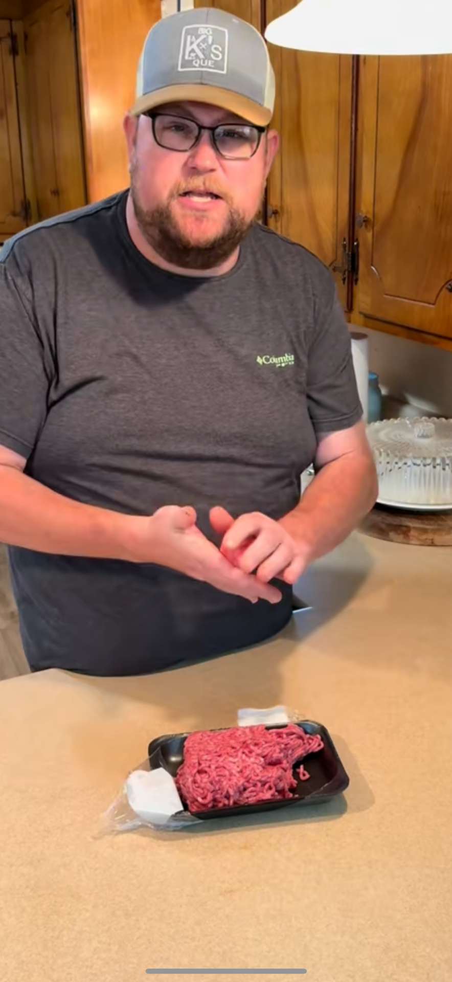 A man wearing glasses, a gray T-shirt, and a cap stands in a kitchen, preparing to cook with a package of ground beef on the counter in front of him.